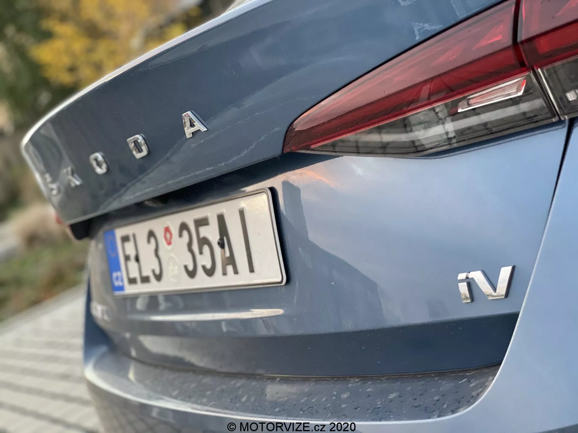 Close-up da seção traseira de um Škoda Octavia Limousine 2020, capturado de um ângulo baixo focando na tampa do porta-malas, parte do para-choque traseiro, com logotipo Škoda em cromo, emblema iV para variante híbrida ou elétrica, placa de licença em estilo europeu, luzes traseiras estreitas e horizontais que se estendem até a tampa do porta-malas, e uma faixa refletora no para-choque traseiro.