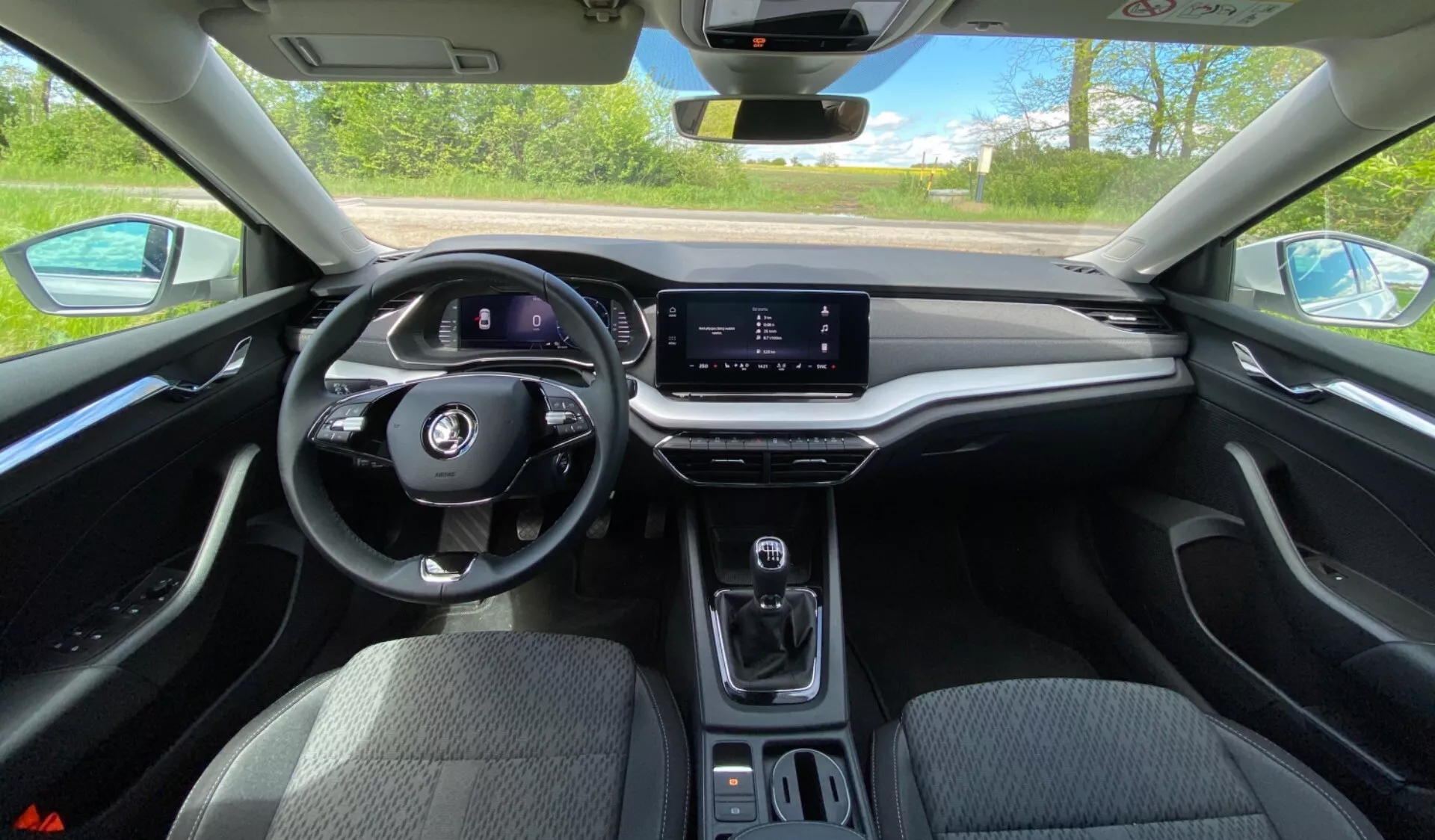 Vista interior do Skoda Octavia 2020 a partir da perspetiva do condutor, mostrando o volante multifuncional, o painel de instrumentos digital, o ecrã tátil de infoentretenimento orientado horizontalmente, a consola central minimalista com seletor de velocidades, suportes para copos e painel de toque suave com acabamento em imitação de madeira em condições de luz diurna brilhante.