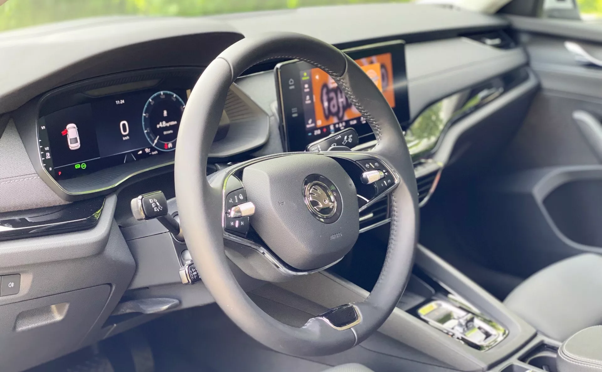 Vista interior de um veículo moderno focando no painel do lado do motorista a partir de uma perspectiva ligeiramente à direita do banco do motorista. A imagem destaca claramente um volante de três raios com botões de controle, um painel de instrumentos totalmente digital exibindo a velocidade do veículo e outras informações vitais, e uma tela central de infoentretenimento de alta resolução mostrando o sistema de controle climático do carro. Materiais de toque suave de alta qualidade e acabamentos visíveis no painel e na porta realçam a sensação de luxo no interior do carro. O primeiro plano borrado sugere a presença de um assento para passageiro.