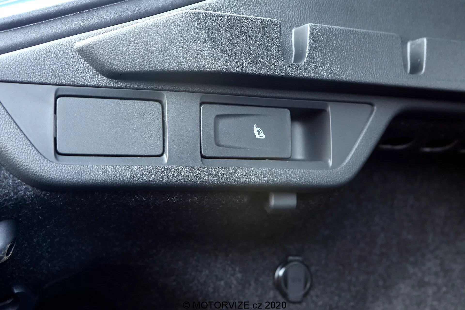 A imagem captura uma vista detalhada do painel de controle de um carro a partir de uma perspectiva ligeiramente inclinada de cima. O painel apresenta dois botões retangulares embutidos em uma moldura de plástico cinza texturizado. O botão à esquerda está sem marcação, enquanto o botão à direita possui um ícone de uma tampa de porta-malas entreaberta, indicando uma função de abertura do porta-malas. Uma pequena luz indicadora, provavelmente um LED, está situada à direita dos botões. Abaixo dos botões há um objeto cilíndrico, possivelmente uma abertura manual ou de emergência do porta-malas. A foto não revela o contexto mais amplo do interior do carro.