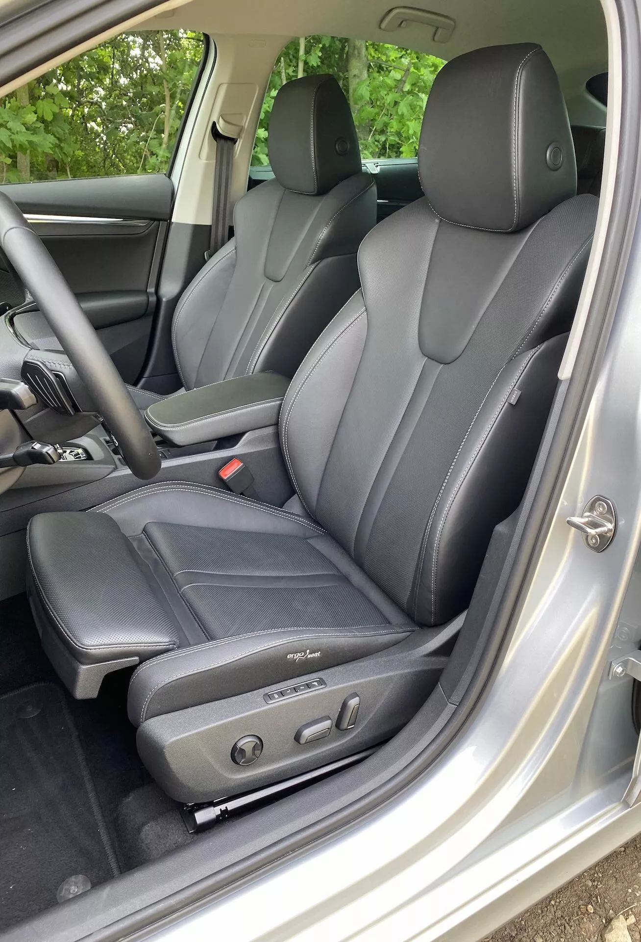 Vista interior do Škoda Octavia limousine 2020 focada na área do condutor, com bancos de couro escuro de design ergonómico e controles de ajuste elétrico, vista do lado esquerdo através da porta do condutor aberta. Os apoios de cabeça visíveis para o condutor e passageiros traseiros indicam capacidade para pelo menos quatro ocupantes. O acabamento interior em prata ou cinza claro contrasta com os bancos, e o painel da porta inclui controles de janelas e espelhos elétricos, destacando o conforto e luxo tecnológico.