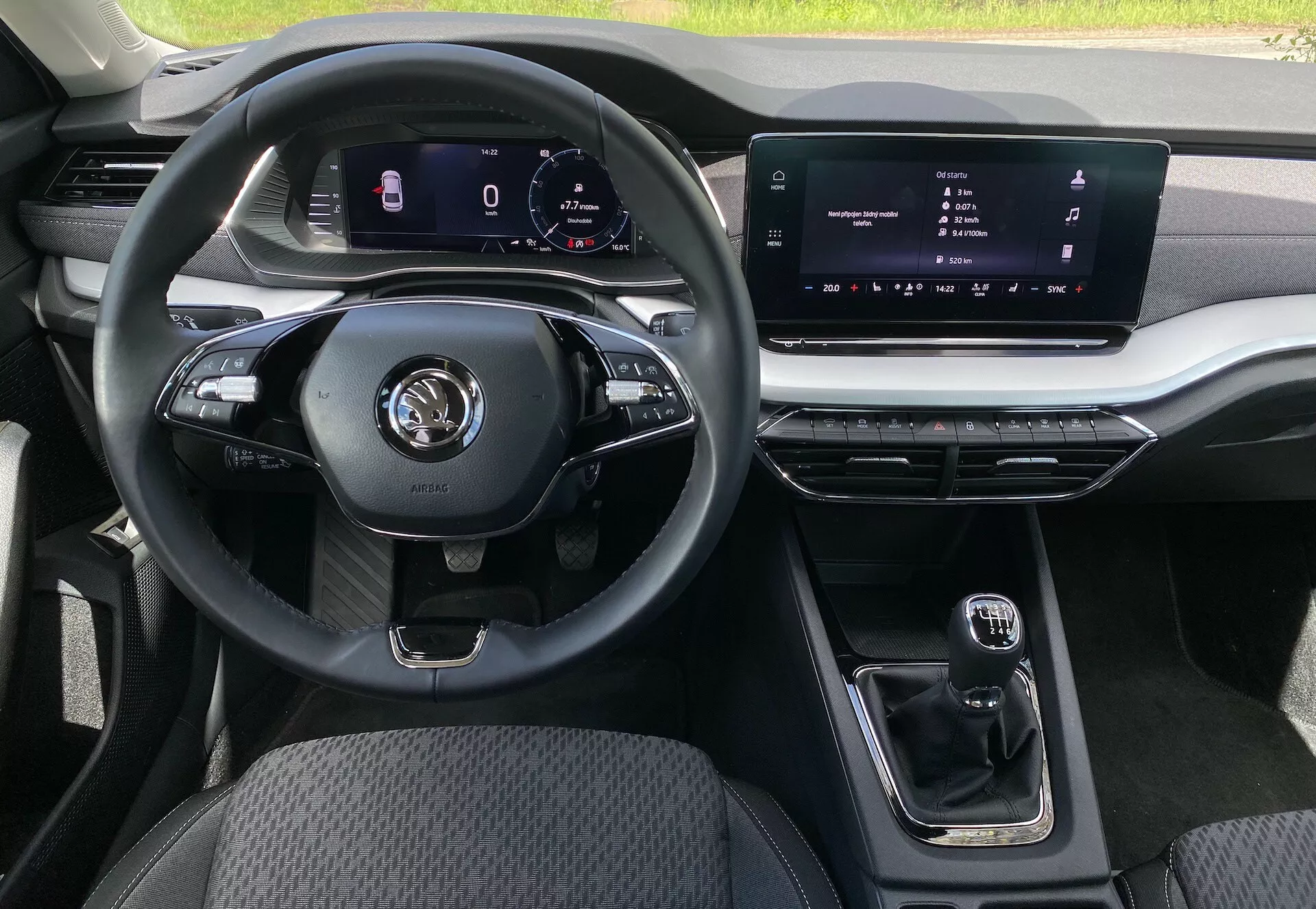 Vista interior do sedan Škoda Octavia 2020 da perspetiva do assento do passageiro, destacando um volante multifuncional de três raios com vários controles para média e chamadas em modo mãos-livres, um painel de instrumentos digital para navegação e estatísticas do veículo, um ecrã central tátil de infotainment, uma alavanca de mudanças automática compacta, acentos metálicos nas saídas de ar e no sistema de infotainment, estofos de tecido durável no assento do motorista e iluminação ambiente personalizável.