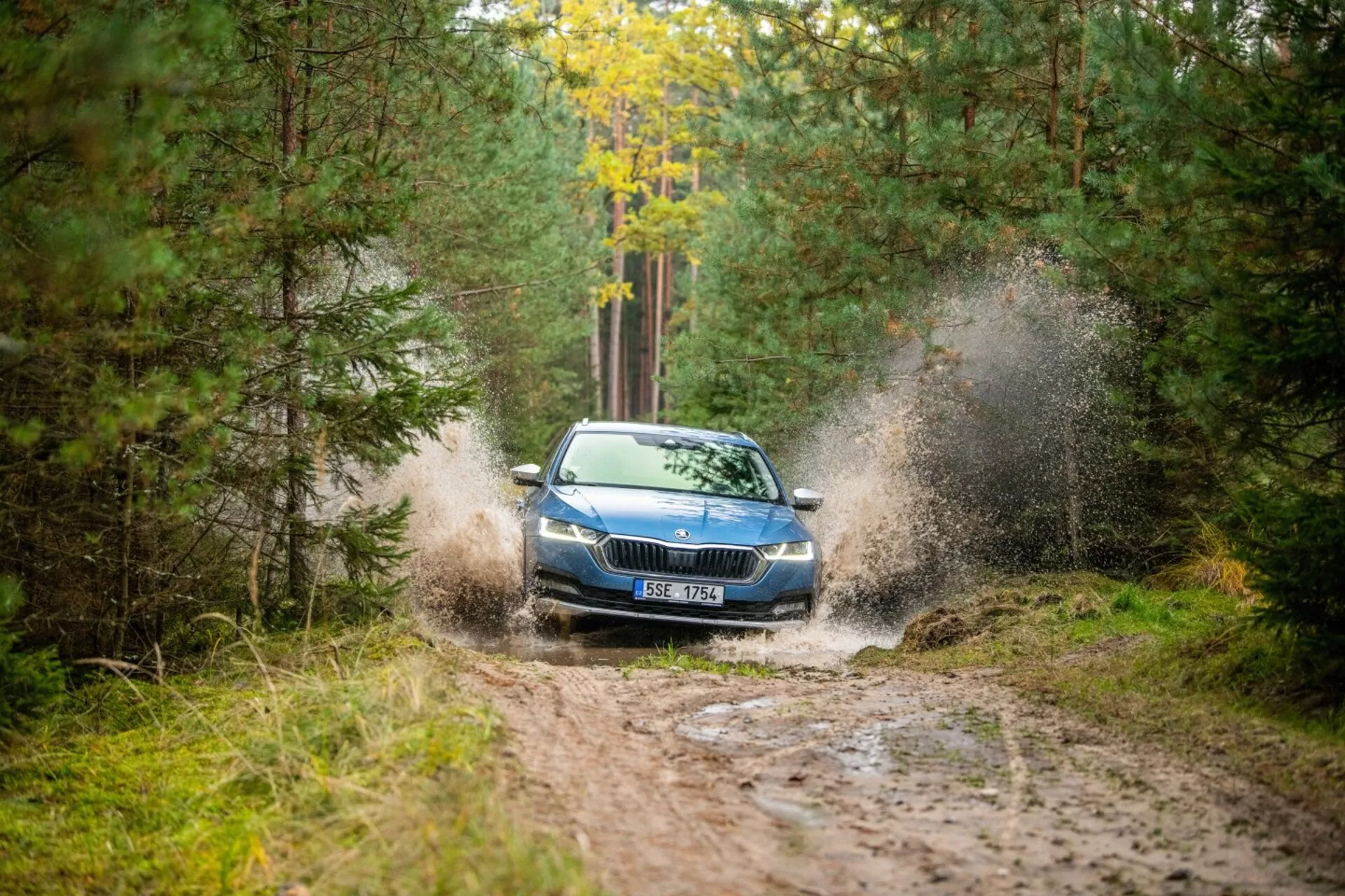 Limousine Skoda Octavia de 2017 dirigindo através de uma poça de lama em uma área florestal, destacando sua criação dinâmica de respingos, grade frontal, faróis e capô com uma visão parcial do teto de um ângulo frontal direito.