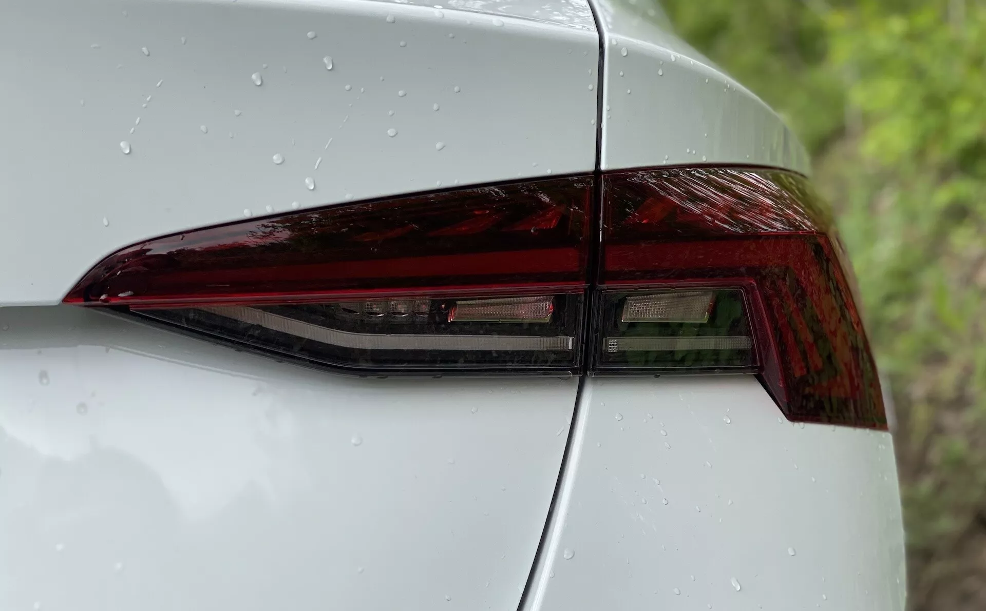 Close-up da parte traseira esquerda de um Škoda Octavia 2020 com foco no conjunto de luzes traseiras. A imagem é tirada de um ângulo diagonal que destaca o design contemporâneo das luzes LED com ângulos acentuados e acabamento brilhante. Parte do painel traseiro quarto e da tampa do porta-malas são visíveis, mostrando uma integração perfeita e uma estética aerodinâmica. A superfície lisa tem gotas de água, indicando precipitação recente ou uma lavagem recente.