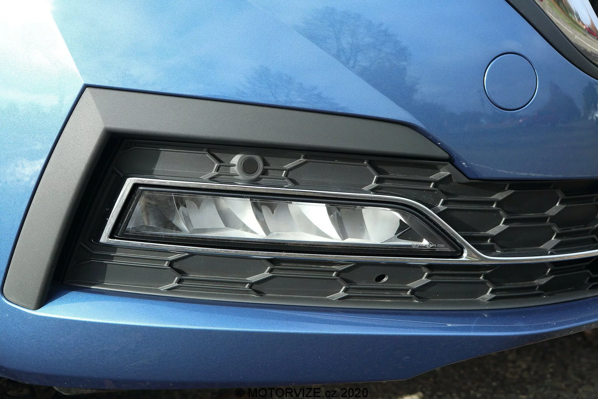 Detalhe em close-up da frente do Skoda Octavia 2020 (Octavia Limousine), enfatizando o farol esquerdo e a grade. O farol LED moderno com design angular acentuado está situado em uma reentrância esculpida, fundindo-se perfeitamente com a carroceria azul metálico do carro. A grade adjacente tem um padrão hexagonal com plástico preto e acentos cromados, refletindo o estilo automotivo contemporâneo.