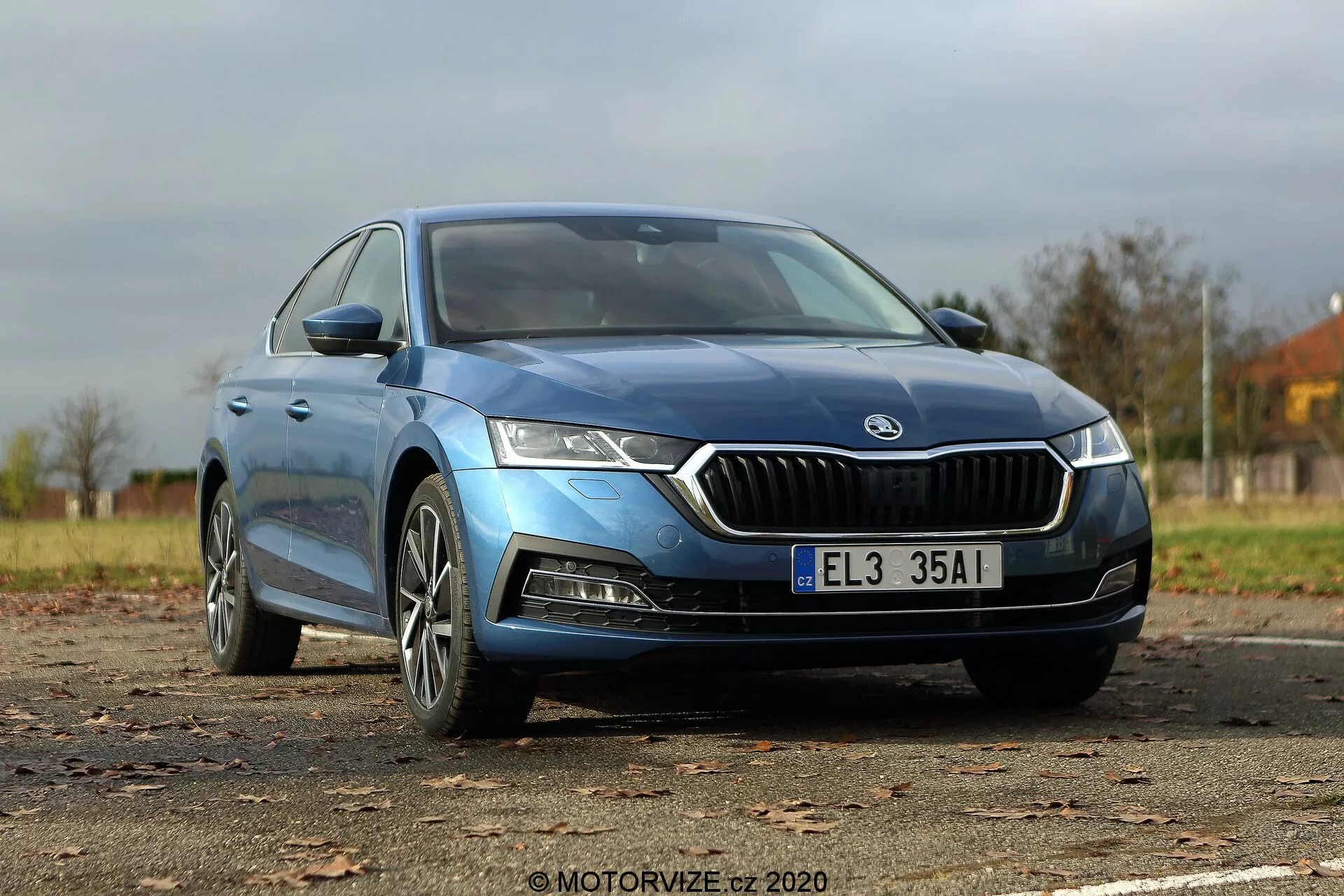 Imagem de um Škoda Octavia Limousine azul 2020 capturada de um ângulo frontal baixo de três quartos, destacando a distinta grade frontal com contorno cromado, faróis LED afiados, capô contornado, rodas de liga modernas e espelhos laterais elegantes com sinais de seta integrados. A carroceria suave e as janelas ligeiramente escurecidas sem moldura indicam um nível de acabamento superior, enquanto todo o comprimento do carro é exibido, incluindo os emblemas visíveis que indicam o modelo ou tipo de motor.