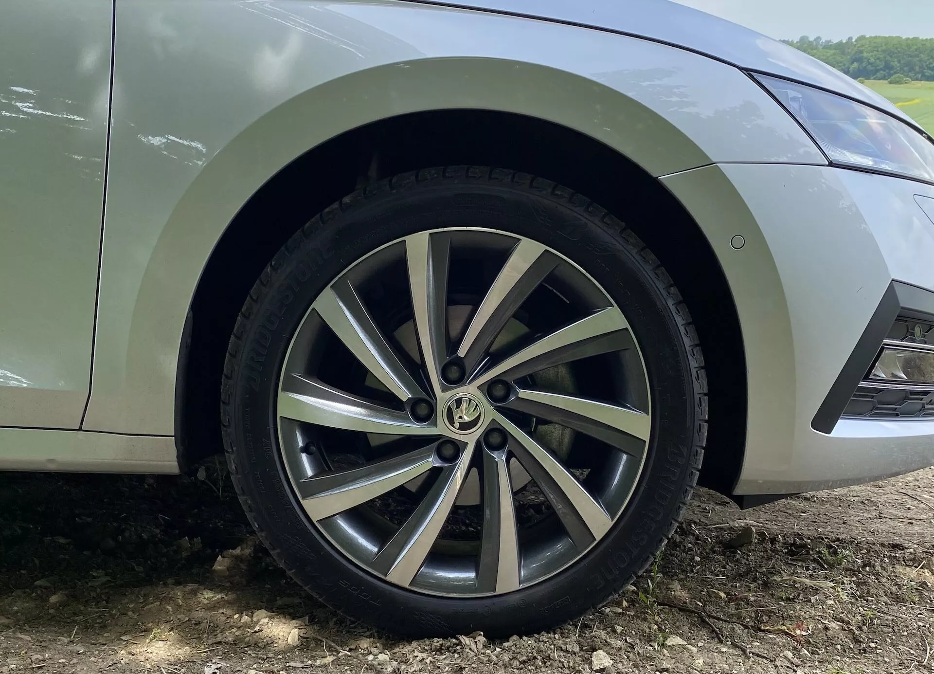 Visão aproximada da roda dianteira e do para-lama inferior de um Skoda Octavia Limousine 2020, destacando o design da roda de liga leve com múltiplos raios, com uma mistura de superfícies prateadas pintadas e usinadas. O sulco do pneu é visível e mostra pouco desgaste. O carro está estacionado em uma superfície de terra com respingos de lama visíveis atrás da roda, indicando uso recente fora de estrada ou em estrada não pavimentada. Apenas a seção do carro desde a porta dianteira inferior até o para-choque dianteiro é mostrada em um ângulo baixo, capturando os detalhes da roda e do para-lama.