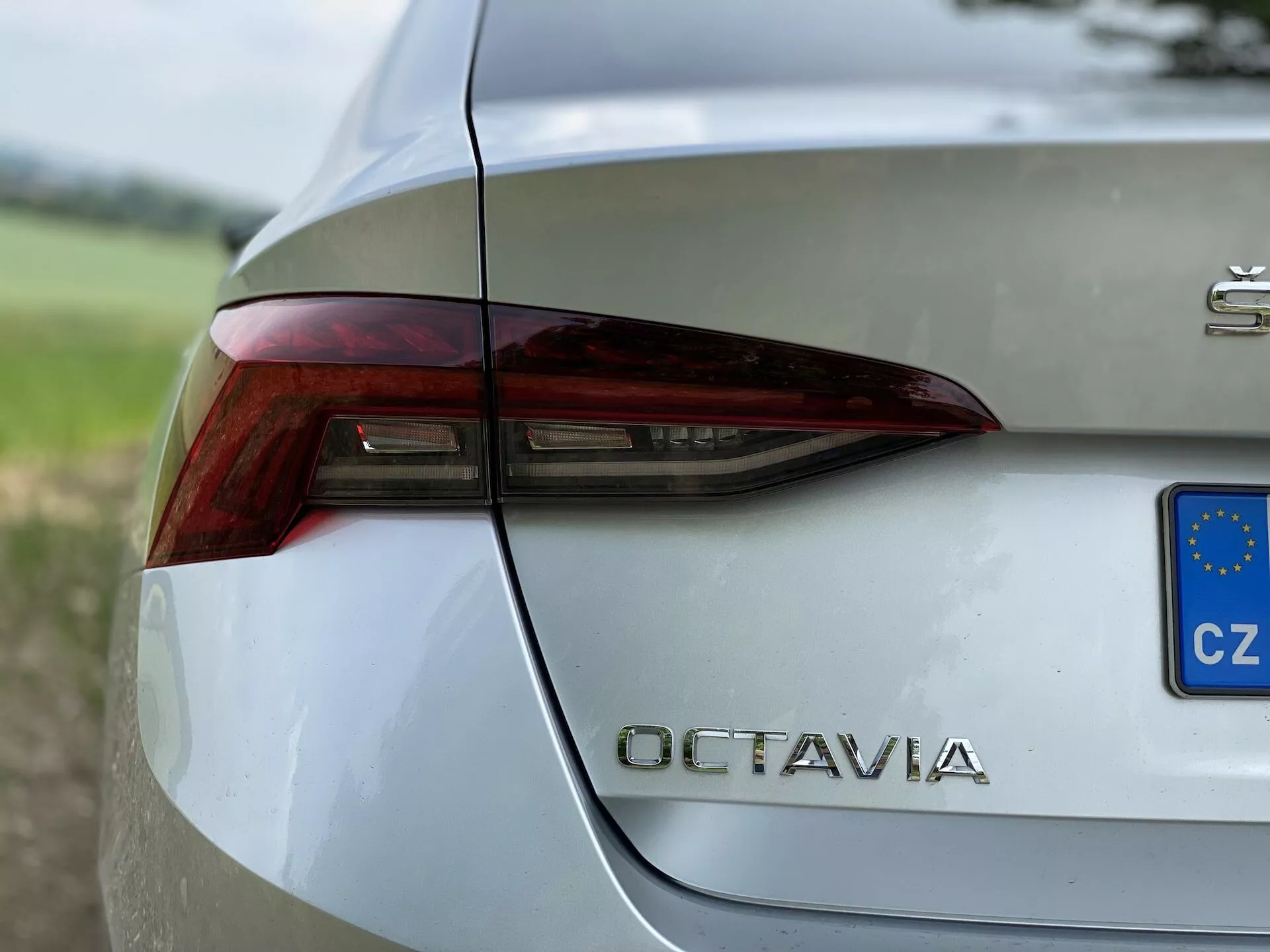 Vista de perto da seção traseira direita de um Škoda Octavia 2020 prateado. A imagem captura o design nítido e angular da luz traseira com elementos integrados para luzes de freio, indicadores de direção e possivelmente luzes de ré. A tampa do porta-malas exibe de forma destacada a designação do modelo 'OCTAVIA' em letras cromadas abaixo da luz traseira. Um pequeno retângulo azul e amarelo com estrelas indica o registro da República Checa. A carroceria metálica polida do veículo reflete o ambiente ao redor, com um fundo desfocado de vegetação, destacando seu design sofisticado.