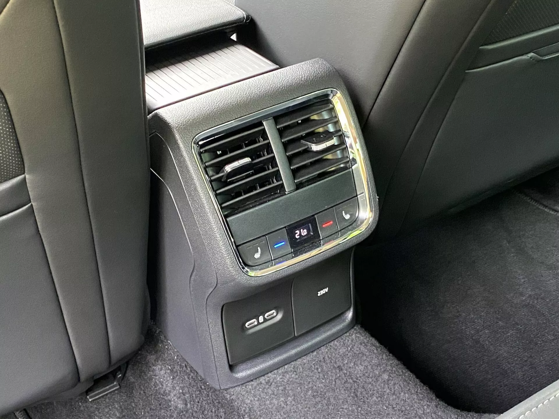 Vista detalhada do console de controle de clima traseiro na parte de trás de um Skoda Octavia limousine 2020. Capturada de um ângulo fora do centro, oferecendo uma visão clara do painel central localizado na parte de trás do apoio de braço dianteiro. Apresenta botões de controle climático digital com ajustes de temperatura, entradas de ar verticais, um display digital de temperatura ajustado em 21℃, portas USB para recarga e possível funcionalidade de aquecimento ou resfriamento dos assentos traseiros. Os materiais são uma combinação de plástico e tecido, em um esquema de cores preto e cinza escuro sob iluminação natural.