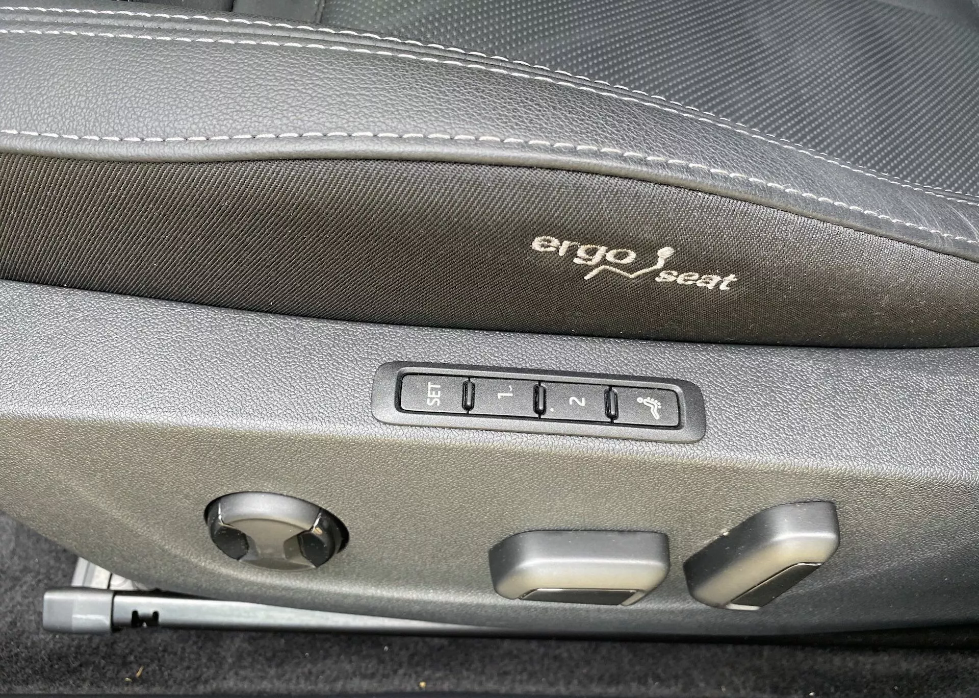 Close-up do interior do Škoda Octavia 2020, focado nos controles de ajuste ergonômico do assento. 'Ergo seat' está bordado na parte superior escura do assento. Abaixo do bordado, cinco botões são visíveis: um para configurações de memória do assento e quatro para ajustes do assento como para frente, para trás, para cima, para baixo e inclinação do encosto. Há também um botão redondo, provavelmente para ajustar o suporte lombar ou outros ajustes multifuncionais do assento, indicando conforto superior e tecnologia avançada neste veículo.