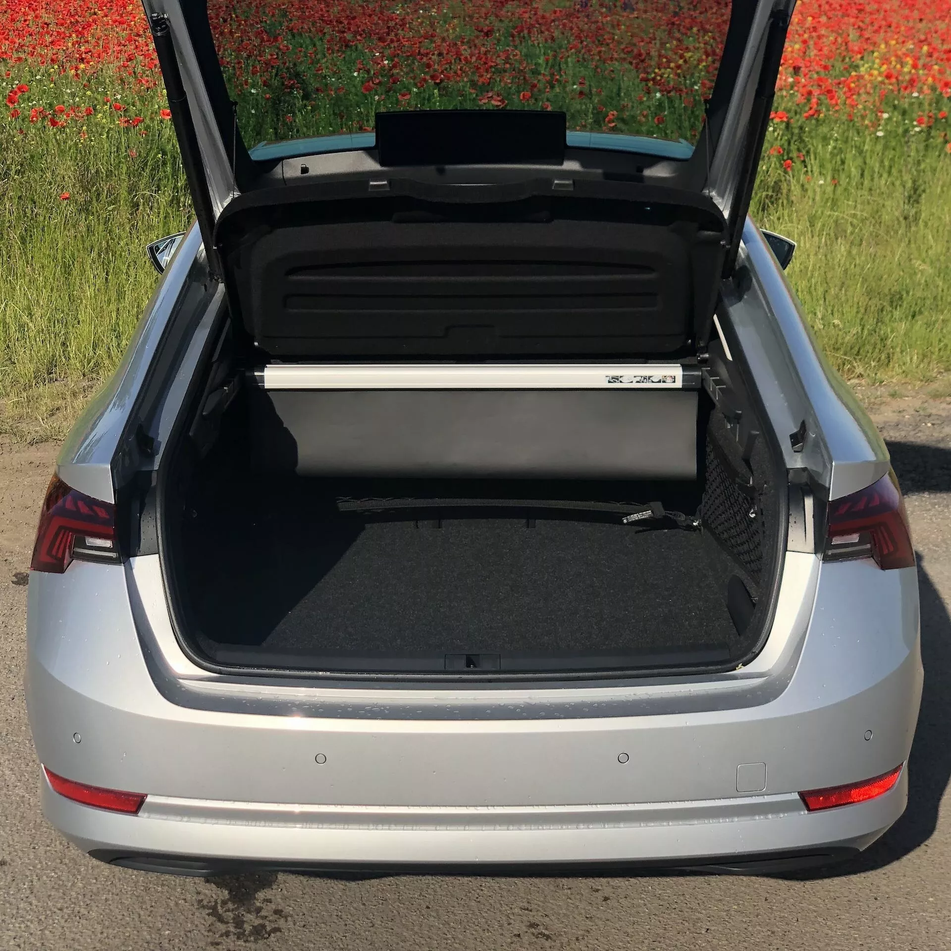 Vista de ângulo alto de um sedã Skoda Octavia 2020 prateado com o porta-malas aberto, mostrando a área de carga espaçosa e organizada. O para-choque traseiro, os distintivos conjuntos de luzes traseiras vermelhas e parte do emblema do carro são visíveis, com um campo de flores vermelhas ao fundo.