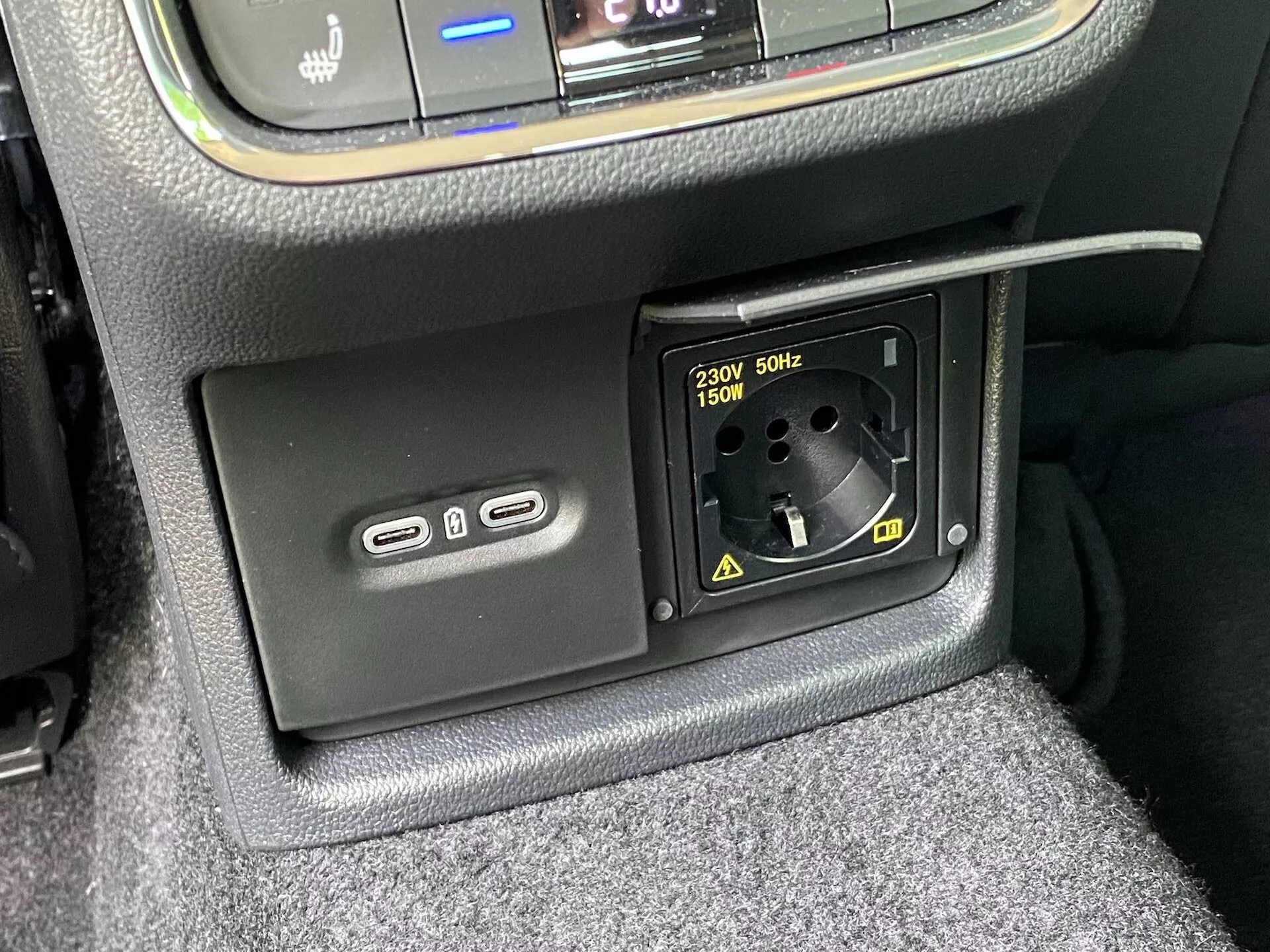 Close-up das características de alimentação em um interior do Skoda Octavia 2020, mostrando a parte inferior da consola central com duas portas USB-A à esquerda para carregamento ou ligação multimédia e uma tomada de corrente de 230V AC para fichas europeias com suporte até 150W à direita, cercada por acabamento texturizado e carpete visível.