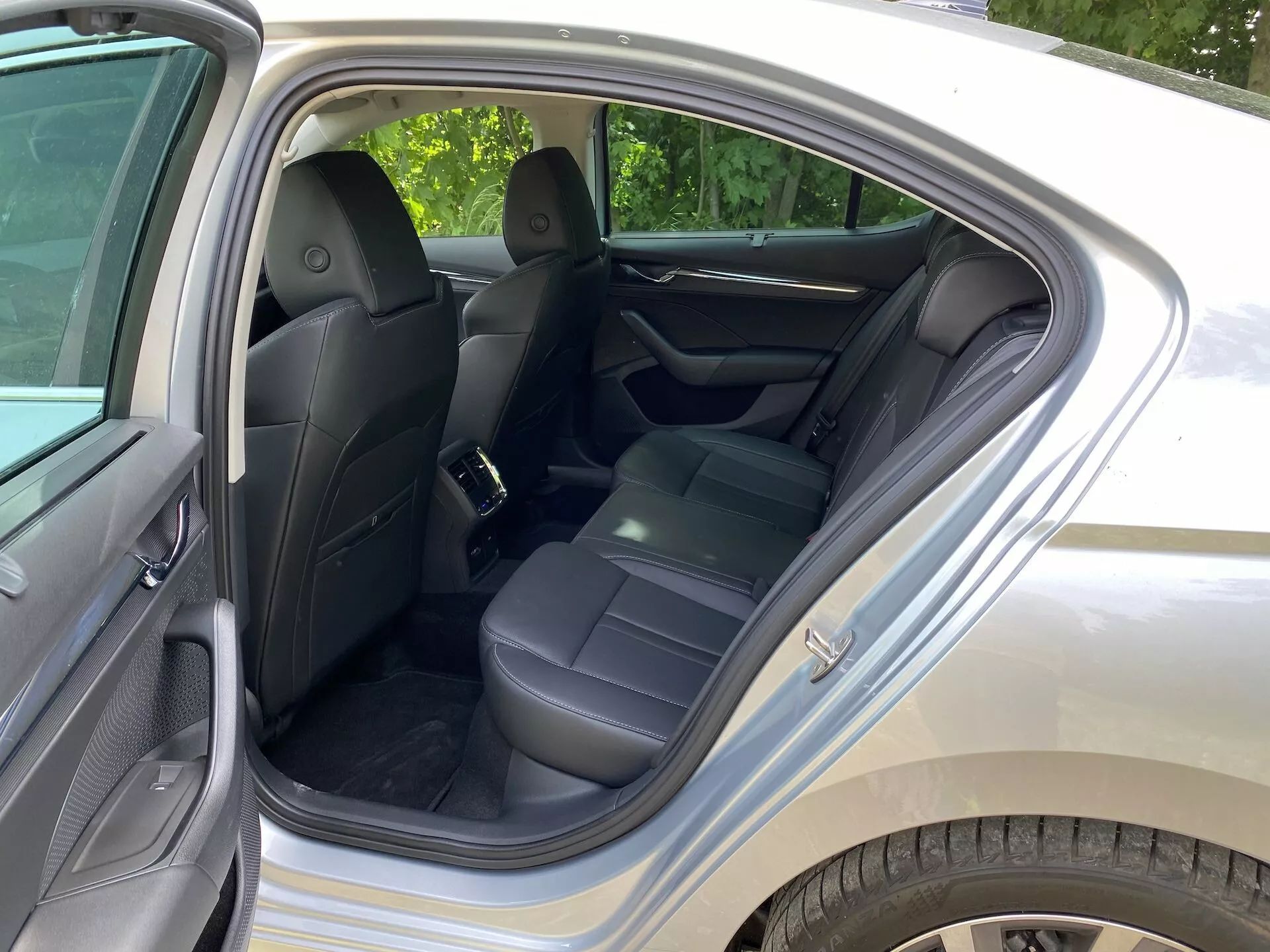 Imagem do interior do Skoda Octavia 2020, focada no compartimento traseiro de passageiros. A porta traseira está aberta, destacando os bancos de couro preto, apoios de cabeça, cintos de segurança e um apoio de braço central dobrado. O painel da porta é feito de material semelhante ao couro para maior conforto. Os destaques visíveis incluem bolsos de armazenamento na parte traseira dos bancos dianteiros, carpete limpo e um ponto de fixação para assento infantil. A iluminação ambiente sugere que a foto foi tirada durante o dia, em uma área suburbana com folhagem verde.