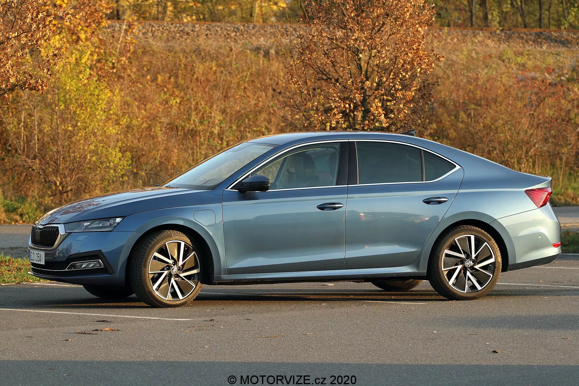 Sedã moderno Skoda Octavia 2020 estacionado ao ar livre, vista lateral em três quartos, mostrando perfil completo, comprimento e altura; detalhes visíveis incluem faróis afiados, linha do teto inclinada, linhas de caráter, janelas com moldura cromada, rodas de liga de múltiplos raios, cenário de folhagem de outono e iluminação natural durante o dia.