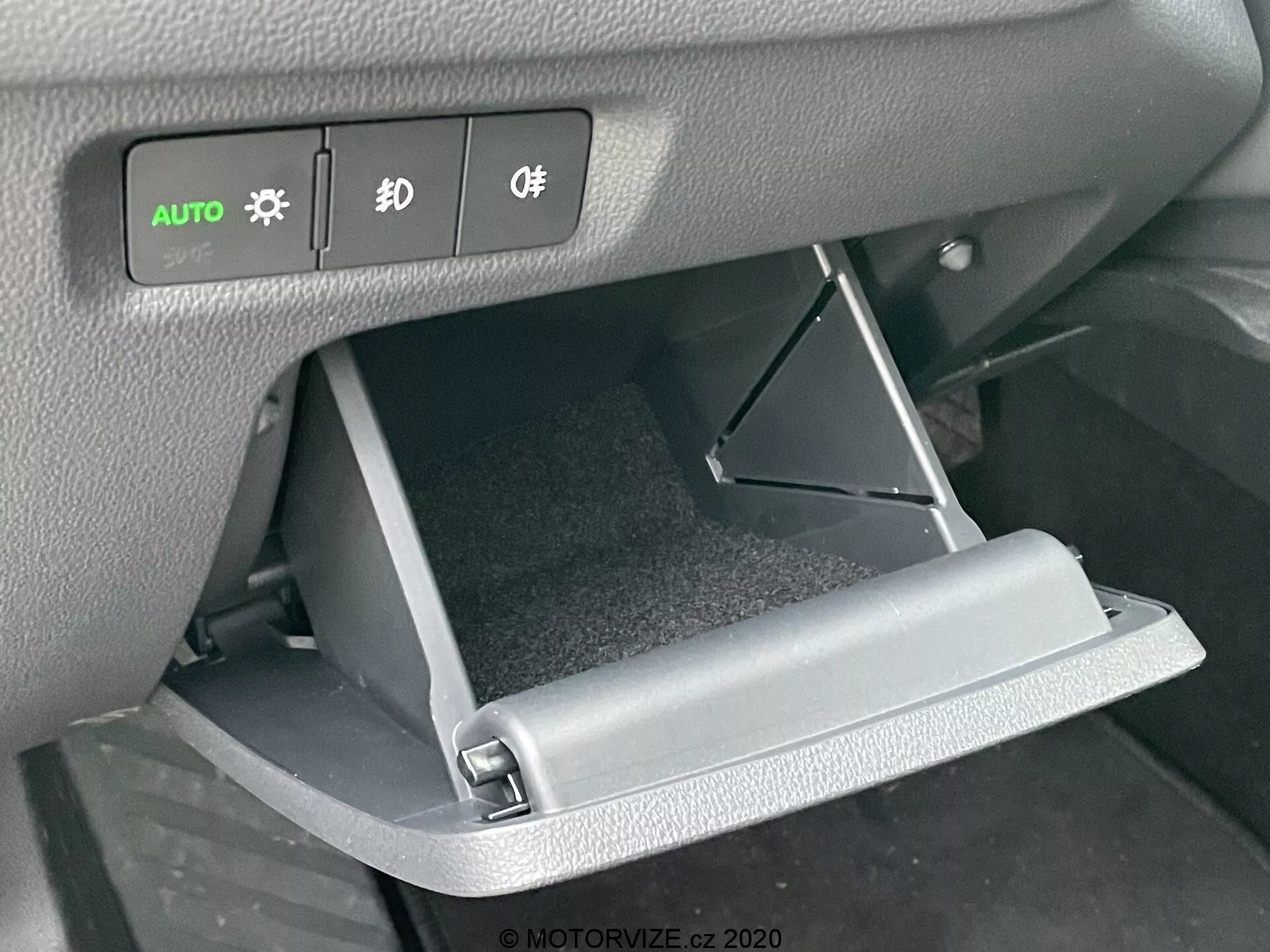 Close-up do interior do Skoda Octavia 2020, focando no porta-luvas aberto e nos controles do painel de instrumentos. O ângulo frontal destaca a porta do porta-luvas aberta para baixo e os botões iluminados dos faróis, luzes de neblina e controles de iluminação acima dela.