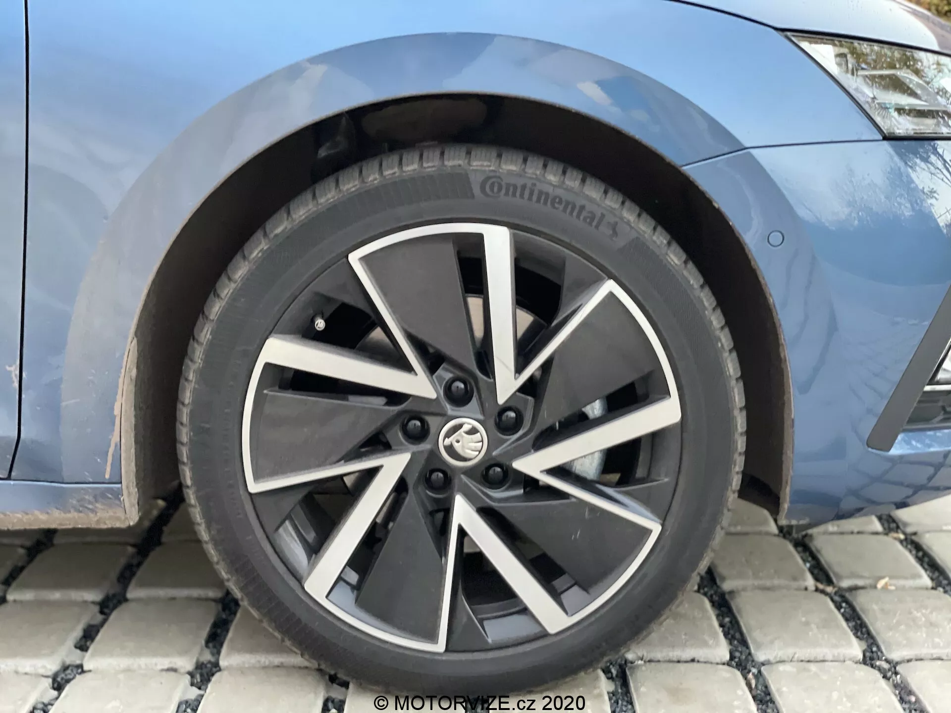 Close-up da roda dianteira e vista parcial do para-lama dianteiro de um Skoda Octavia 2020 limusine. A roda apresenta um design moderno com múltiplos raios em liga de duas cores, predominantemente preto com detalhes prateados nos raios. No centro da roda está o emblema do fabricante. O pneu é da marca Continental, provavelmente da série Conti, com especificações de tamanho alfanuméricas parcialmente visíveis. Atrás dos raios, é possível ver o disco de freio e, possivelmente, a pinça de freio, indicando um sistema de frenagem moderno. A carroceria do carro é mostrada em pintura azul metálica.