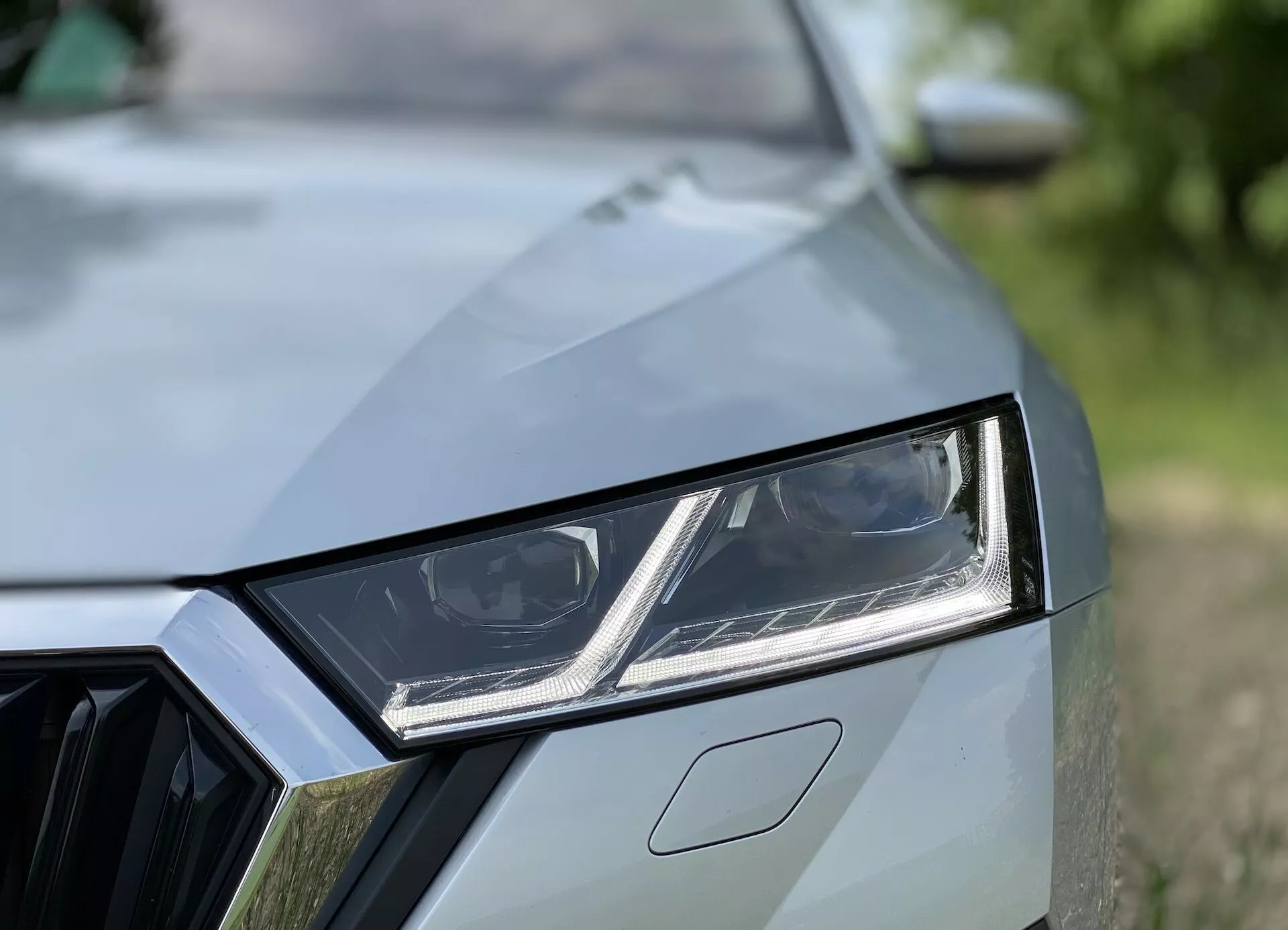 Vista aproximada do lado fronto-esquerdo de um carro moderno mostrando elementos de design detalhados, como o farol LED angular com luzes de rodagem diurna integradas, uma visão parcial da grade com padrão geométrico e o capô com vincos distintivos. O acabamento da pintura de alta qualidade e o fundo desfocado acentuam a estética esportiva e contemporânea do carro.