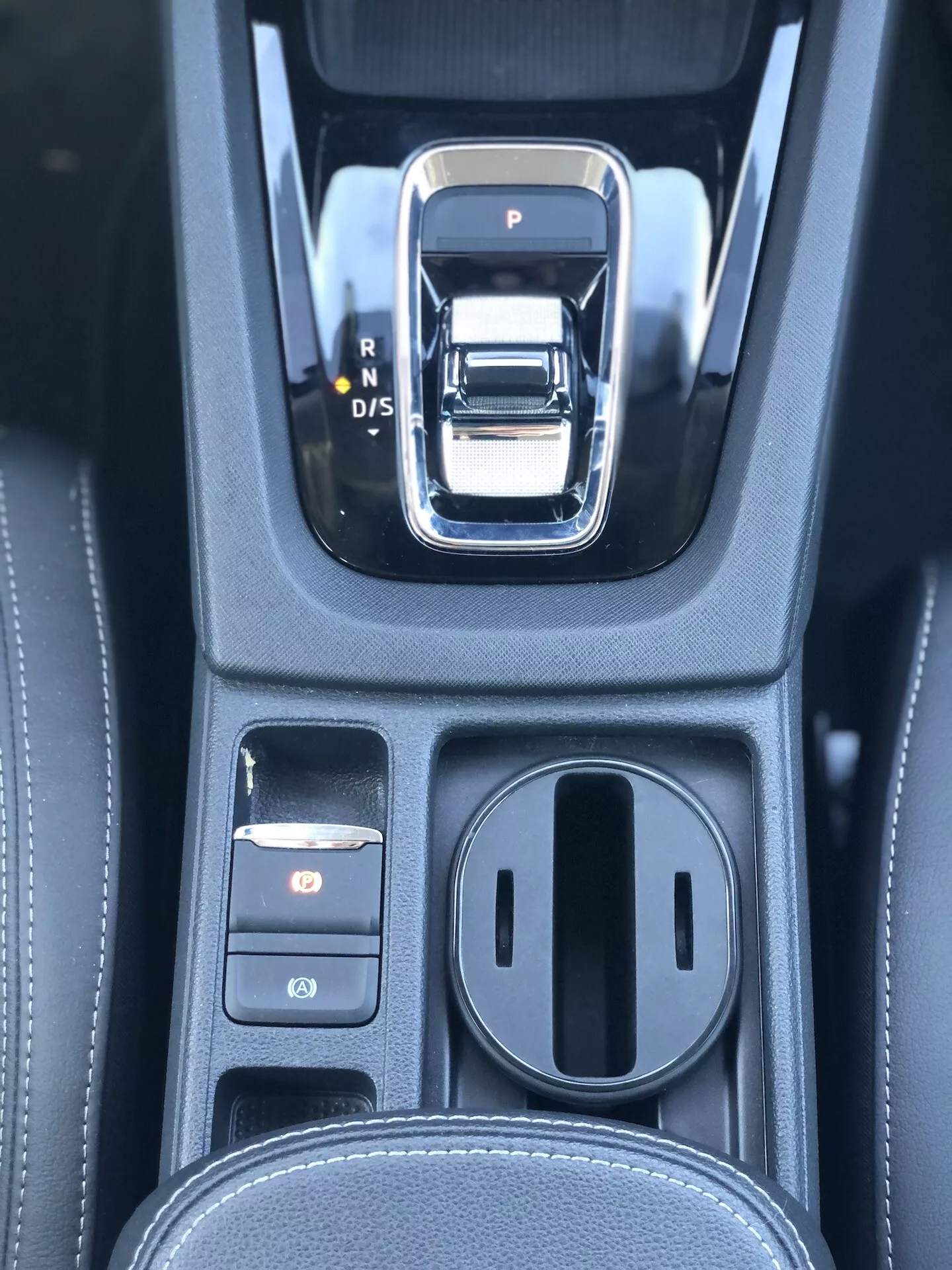Close-up da área do câmbio automático em um Škoda Octavia Limousine 2020. A alavanca de câmbio é uma alavanca eletrônica prateada e preta com opções P, R, N e D/S. À esquerda, há dois botões de controle, com o superior iluminado em vermelho, provavelmente o freio de estacionamento eletrônico. À direita, há um compartimento de armazenamento coberto com dois porta-copos. O console preto apresenta uma textura semelhante à couro com costuras detalhadas, indicando um interior de alta qualidade com sistemas avançados de transmissão e de estacionamento eletrônico.