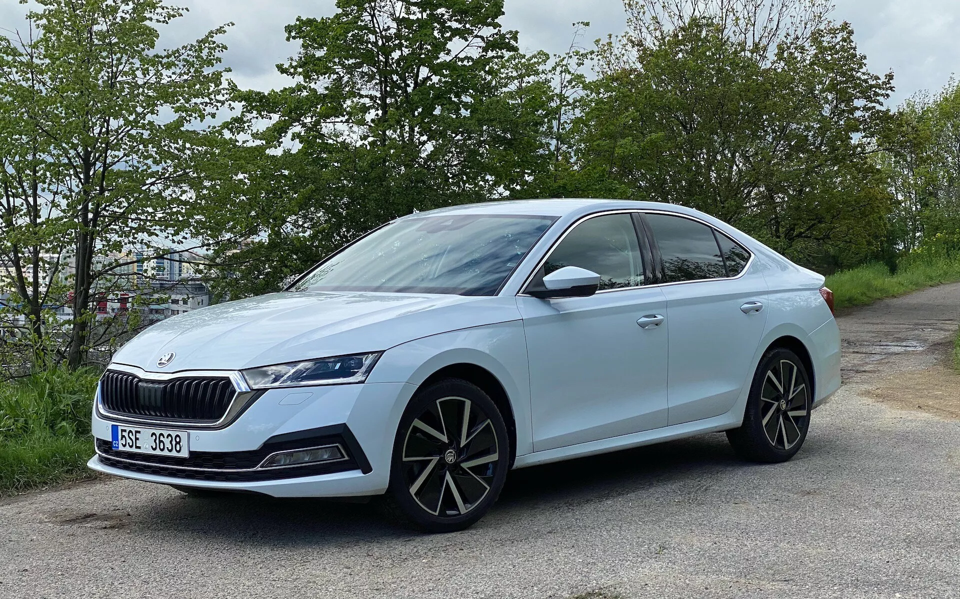 Vista lateral moderadamente angulada de um sedã Skoda Octavia 2020 azul claro, posicionado à beira da estrada com folhagem verde ao fundo. Mostra um design moderno com carroceria de quatro portas, grade proeminente, faróis de LED agressivos, grandes rodas de liga leve multirrayadas, retrovisores da cor da carroceria com indicadores de direção integrados, linha de teto inclinada, janelas escurecidas com moldura cromada, maçanetas embutidas, pneus de perfil baixo e um emblema indicando o modelo ou pacote tecnológico.