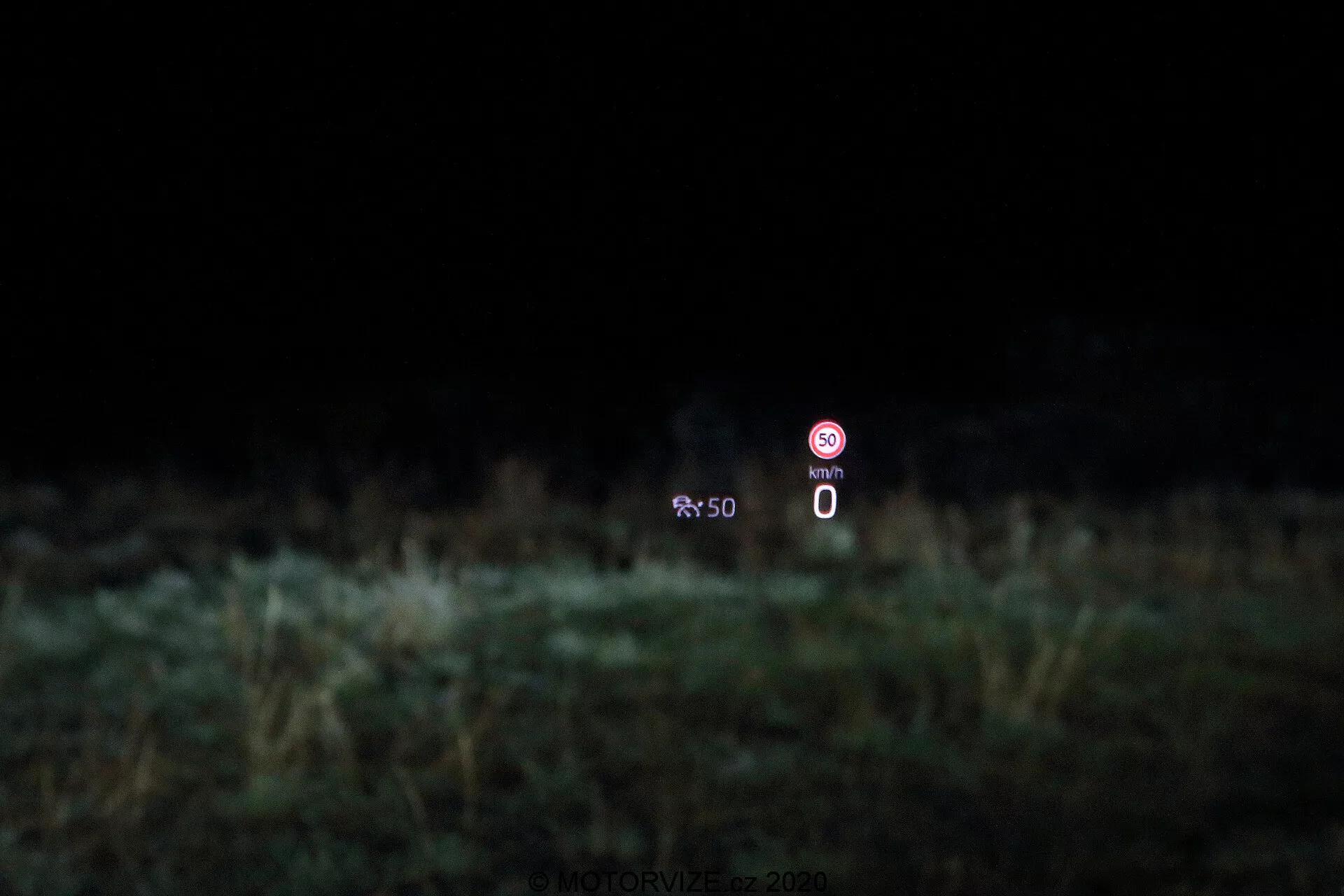 Imagem que mostra a tecnologia head-up display (HUD) em um Škoda Octavia 2020 limousine. O display é projetado no para-brisas, mostrando um velocímetro estático a 0 km/h e um ícone de proibição indicando uma função desativada, com um fundo desfocado de vegetação.