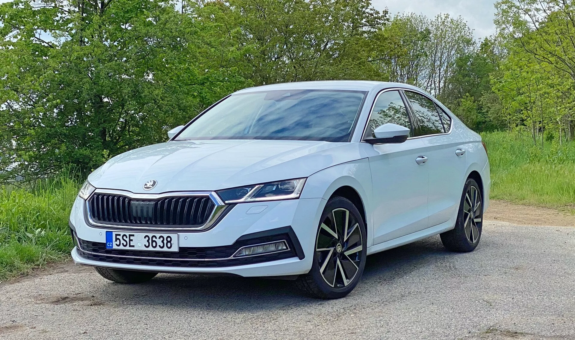 Imagem de um Škoda Octavia 2020 cinza azulado claro posicionado em uma estrada pavimentada com folhagem ao fundo. Tirada em um ângulo de três quartos frontais, a foto destaca detalhes de design: a grade frontal proeminente com lâminas verticais e emblema Škoda, elegantes faróis LED angulares com luzes diurnas, luzes de neblina separadas, grandes rodas de liga leve multi-raios, espelhos laterais com piscas integrados, possíveis sensores de estacionamento frontais e linhas dinâmicas na capota e nos lados. A cena sugere um ambiente diurno sereno com iluminação natural.