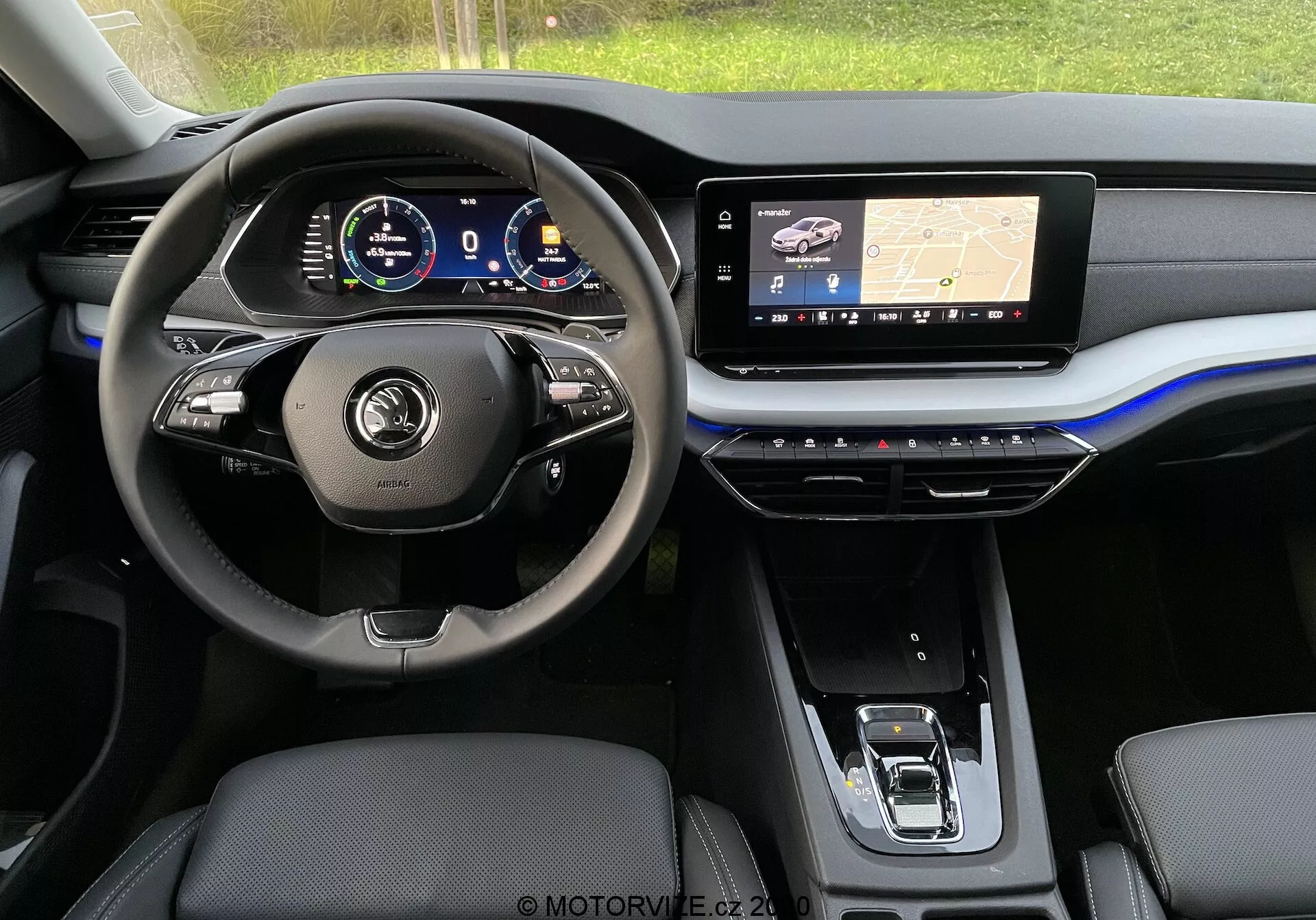 Vista interior de um moderno Skoda Octavia Limousine 2020 a partir do banco traseiro, mostrando o painel frontal e focando na área do assento do motorista. O volante possui botões multifuncionais e revestimento em couro, atrás dele um painel de instrumentos digital exibe vários medidores do veículo. O painel central abriga um grande ecrã tátil de infotainment que exibe um mapa de navegação com tecnologia GPS, enquanto a consola central abaixo contém botões sensíveis ao toque para controle do clima. Visíveis são os assentos dianteiros revestidos em couro ou um material semelhante e um seletor de marchas eletrônico. A estética é elegante com tons escuros, acentos metálicos e um ousado destaque azul que percorre o painel.