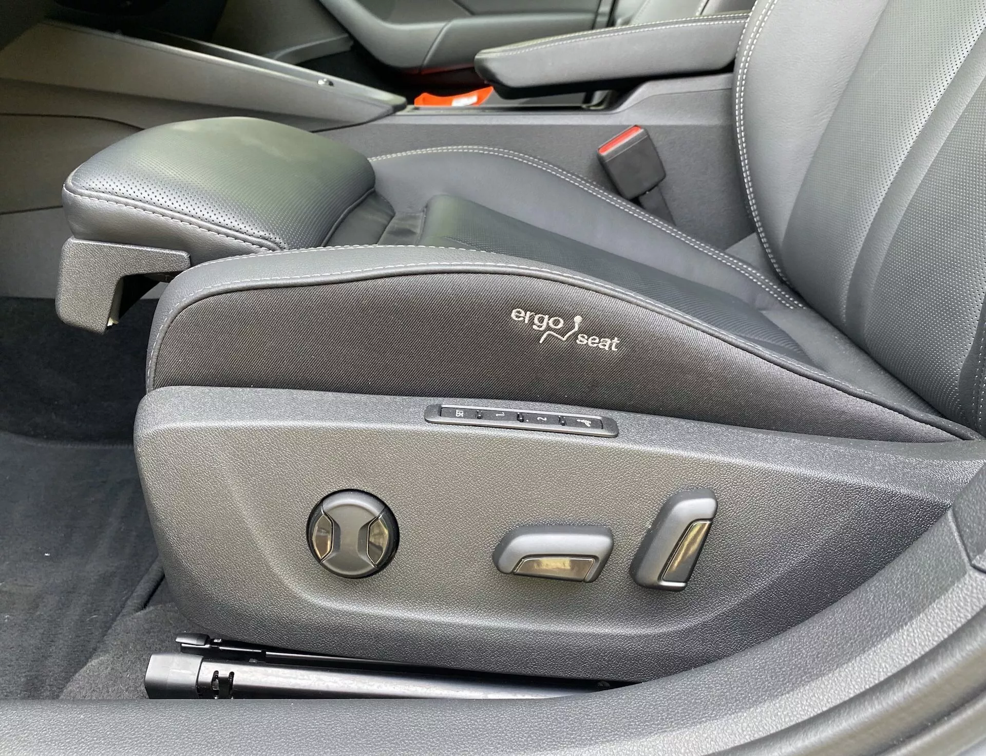 Vista em close-up dos controles laterais do assento do motorista do Skoda Octavia 2020, exibindo design ergonômico com estofamento em tecido escuro, um emblema 'ergo seat' visível e controles de ajuste eletrônico incluindo um ajustador de inclinação circular e dois botões de posicionamento metálicos, destacando o interior premium e tecnologicamente avançado do veículo.