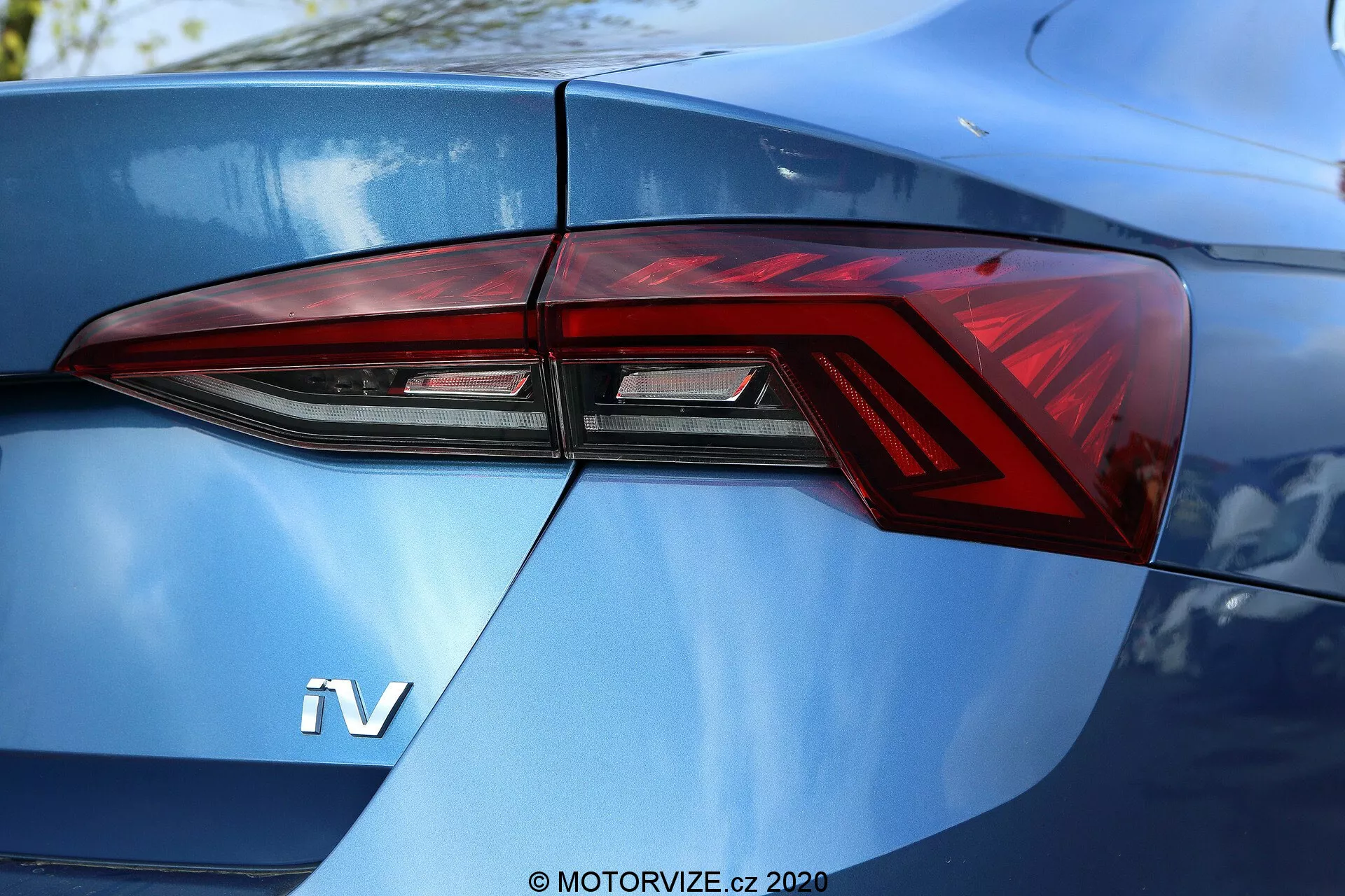Detalhe aproximado da seção traseira de um Skoda Octavia Limousine 2020, focado principalmente no conjunto de faróis traseiros do lado esquerdo visto em um ângulo de três quartos. O design moderno das luzes traseiras apresenta ângulos acentuados e elementos de iluminação LED distintos, integrados de forma impecável nas linhas da carroceria do carro. Um emblema 'iV' visível na tampa do porta-malas azul metálico brilhante sugere um veículo híbrido ou elétrico.