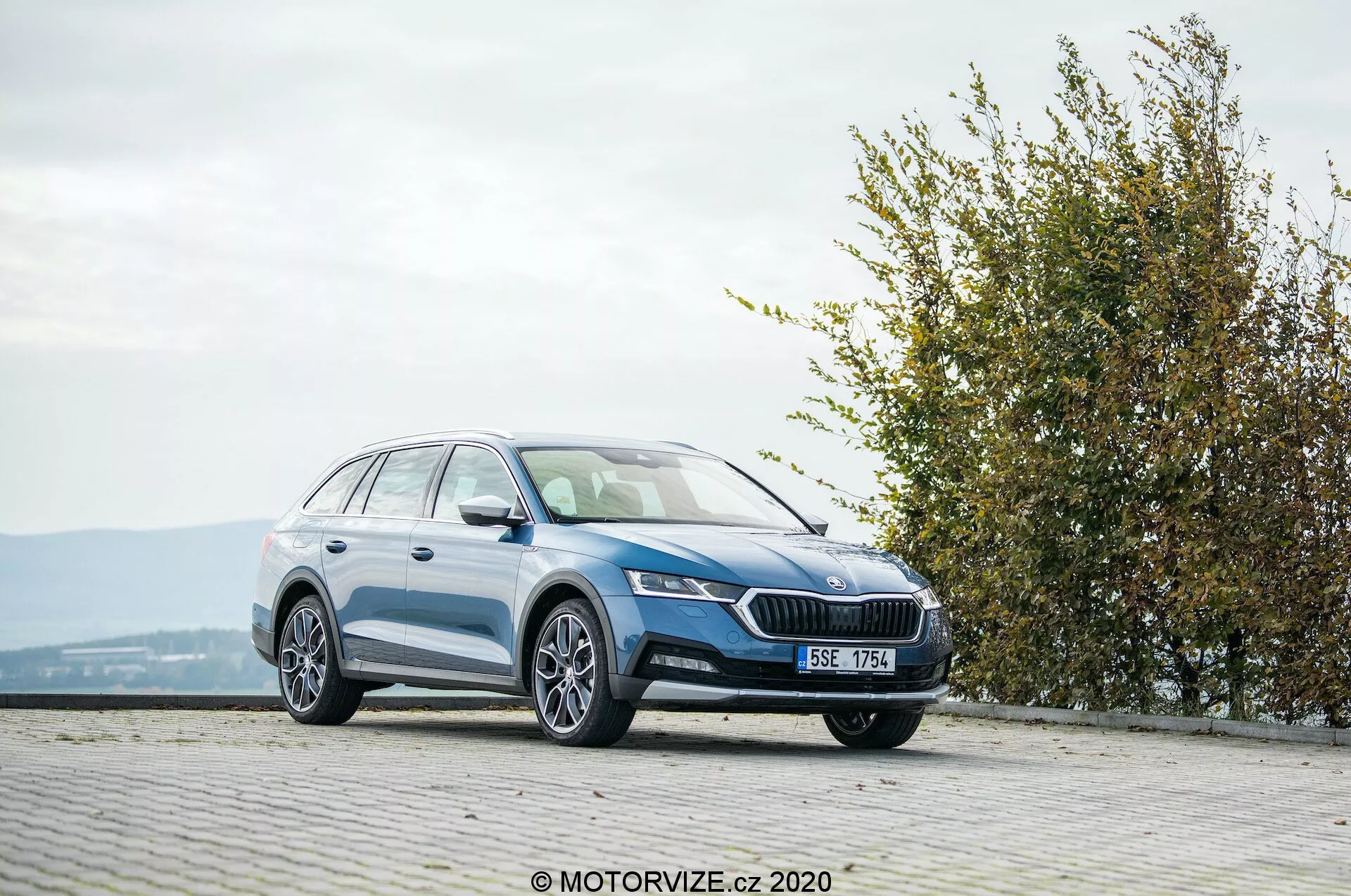 Perua de tamanho completo Skoda Octavia Scout 2020, fotografada de uma vista frontal de três quartos em uma área pavimentada com textura de paralelepípedos em uma localização elevada. A imagem mostra a grade dianteira com um emblema proeminente, faróis angulares elegantes, luzes de neblina integradas, perfil lateral suave com rodas de liga leve, trilhos no teto e um acabamento de pintura brilhante. O contexto circundante inclui uma cerca viva ou arbusto à direita, sugerindo proximidade a um parque ou espaço verde, em condições meteorológicas favoráveis.