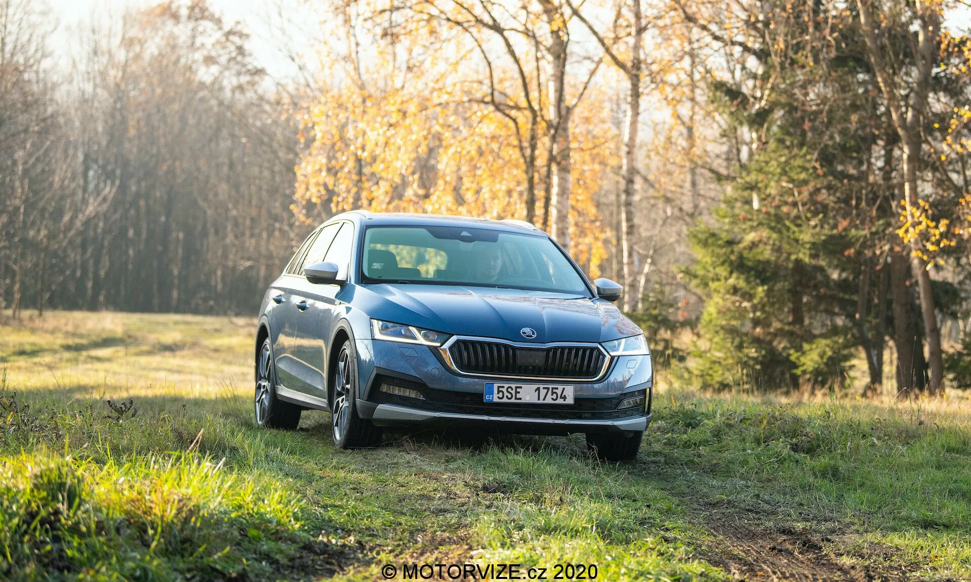 Um sedan Skoda Octavia Scout 2020 azul, fotografado em um ângulo frontal de três quartos, estacionado em uma área gramada com uma floresta ao fundo. A imagem mostra faróis LED modernos, uma grade frontal distinta, luzes de neblina, rodas de múltiplos raios e indicadores de direção integrados nos espelhos laterais. O cenário parece ser de manhã cedo ou no final da tarde com uma luz suave, dando um tom quente ao carro limpo destinado à publicidade ou revisão. O fundo desfocado destaca as características de design do veículo e seu ambiente externo atraente.