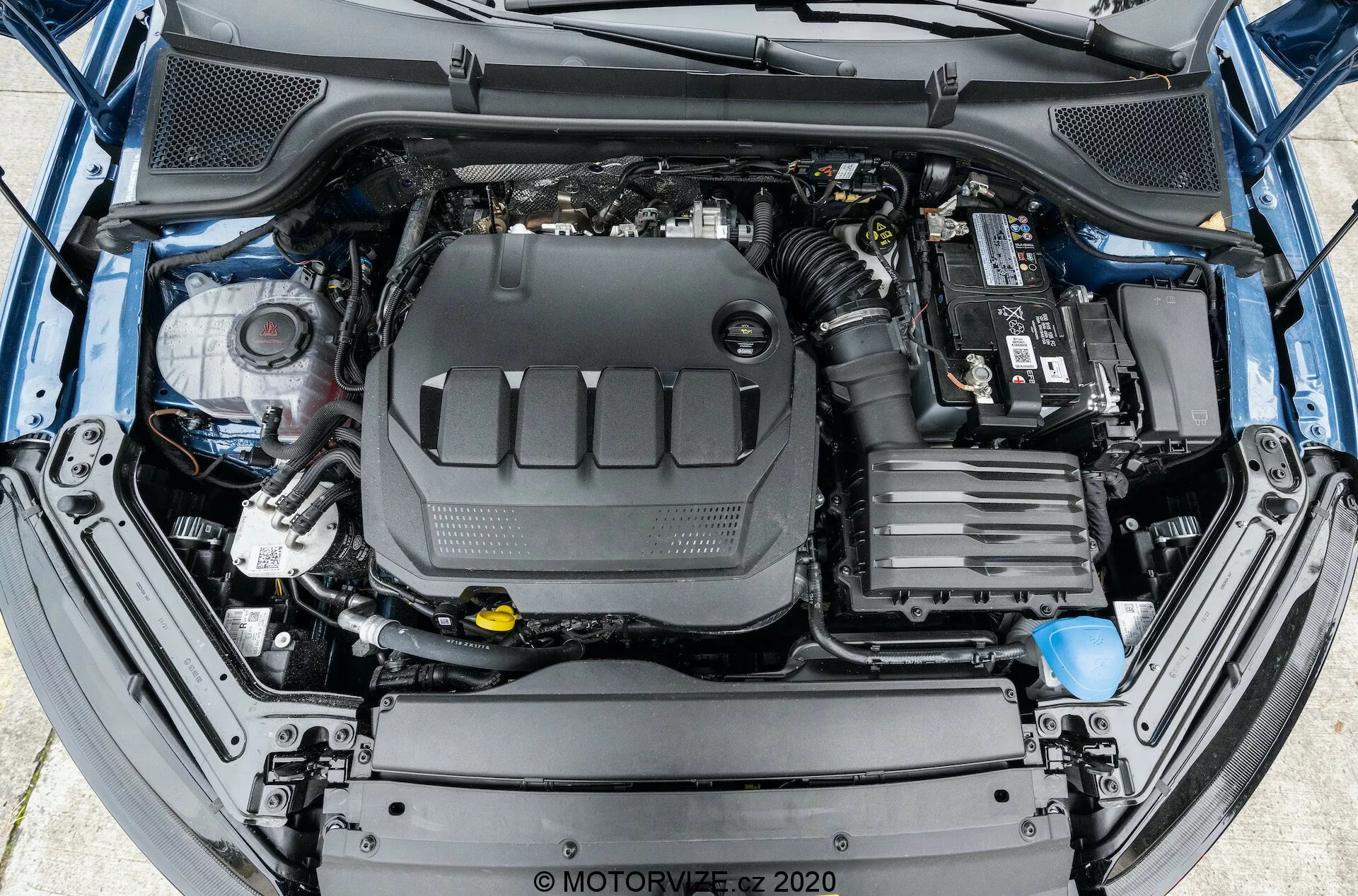 Vista aérea de um compartimento de motor de carro aberto, mostrando uma disposição abrangente do motor e seus componentes circundantes. O bloco do motor está posicionado centralmente, coberto com uma tampa de plástico. À direita, a bateria do carro é visível com conexões de terminal na parte superior. Na parte frontal, são vistos os componentes do sistema de admissão de ar do veículo. No lado esquerdo, há vários reservatórios de fluidos, como o reservatório de fluido de freio com uma tampa amarela. Mangueiras e chicotes de fiação também são visíveis por todo o compartimento do motor. A estrutura do para-choque dianteiro e da grade é parcialmente visível na borda inferior da imagem.