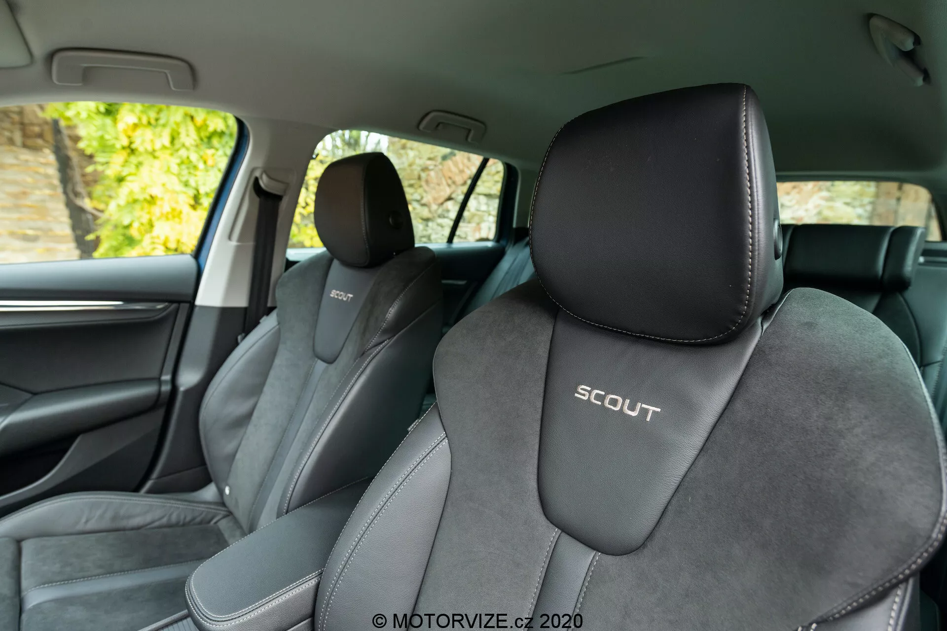 Vista interna de um Skoda Octavia Scout 2020 a partir do banco traseiro, exibindo os assentos dianteiros. O ângulo favorece o lado esquerdo, oferecendo uma vista clara do assento do motorista com um logotipo 'SCOUT' em alto-relevo. Os assentos são estofados em duas tonalidades, predominantemente preto com acentos cinza mais claros e material central semelhante a camurça, destacando um design esportivo. A imagem também apresenta o pilar B, acabamentos interiores escuros, um fragmento da porta dianteira, uma grade de alto-falante e uma maçaneta cromada. O fundo fora das janelas parece mostrar folhagem, indicando que o carro está estacionado ao ar livre com iluminação natural.
