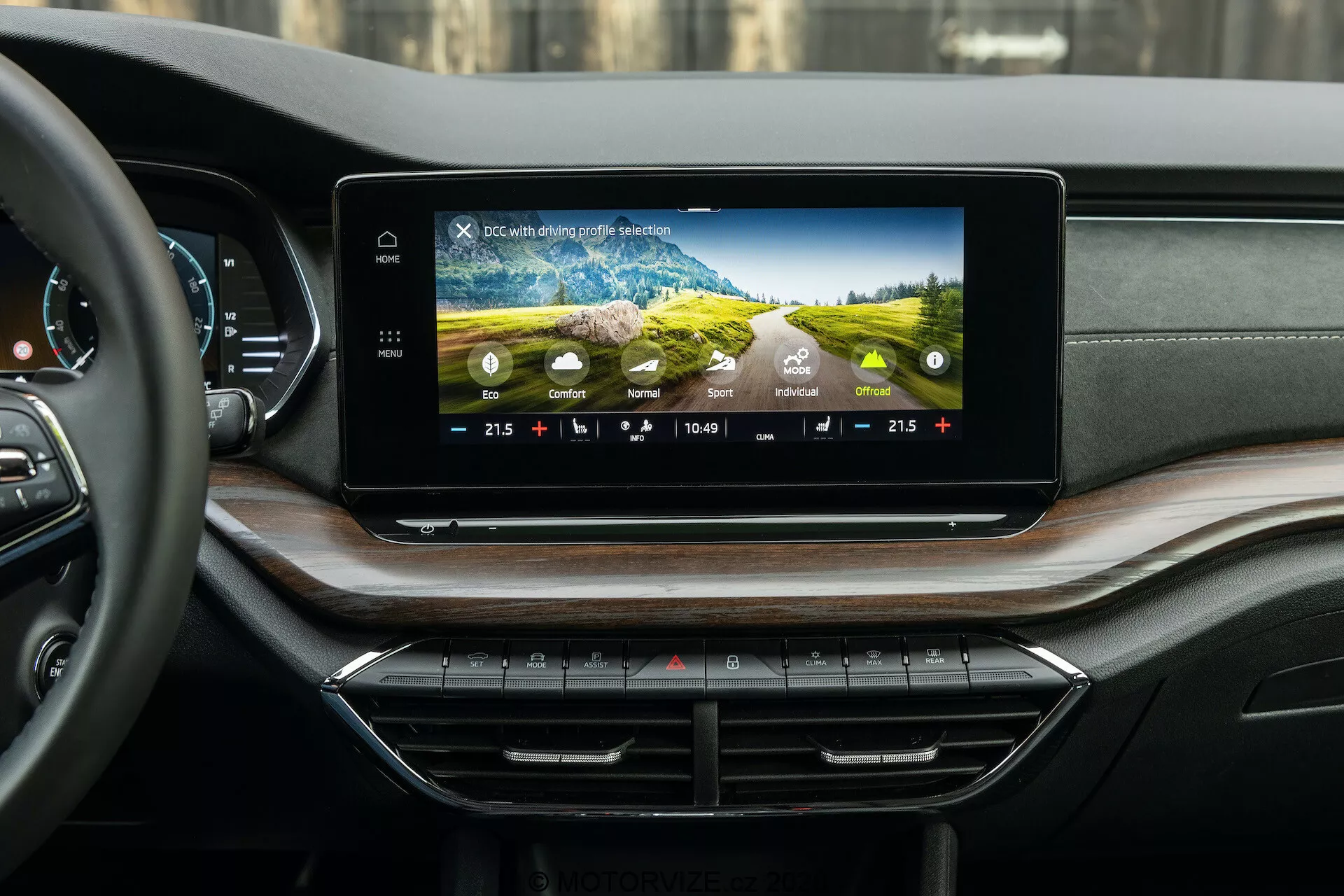 Vista interior do Skoda Octavia 2020 (Octavia Scout) mostrando o moderno sistema de infoentretenimento e painel de instrumentos, fotografado a partir da perspectiva do compartimento traseiro dos passageiros. A tela de toque larga e horizontal no painel de instrumentos exibe gráficos coloridos, configurações de controle de clima, tempo e temperatura externa. Abaixo estão botões e botões de controle de clima ordenadamente dispostos. O painel de instrumentos luxuoso apresenta materiais macios e folheado de madeira, com saídas de ar discretamente integradas.