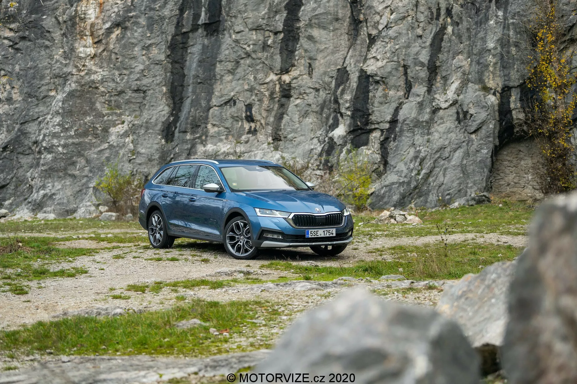 Vista lateral-frontal de um Skoda Octavia Scout 2020 station wagon azul em terreno de cascalho com um fundo rochoso, mostrando faróis LED modernos, grade frontal proeminente, rodas de liga, espelhos laterais, acabamentos de janelas, barras de teto e um acabamento metálico limpo, sugerindo recursos avançados como tecnologia de iluminação, monitoramento da pressão dos pneus e possíveis sistemas de segurança.