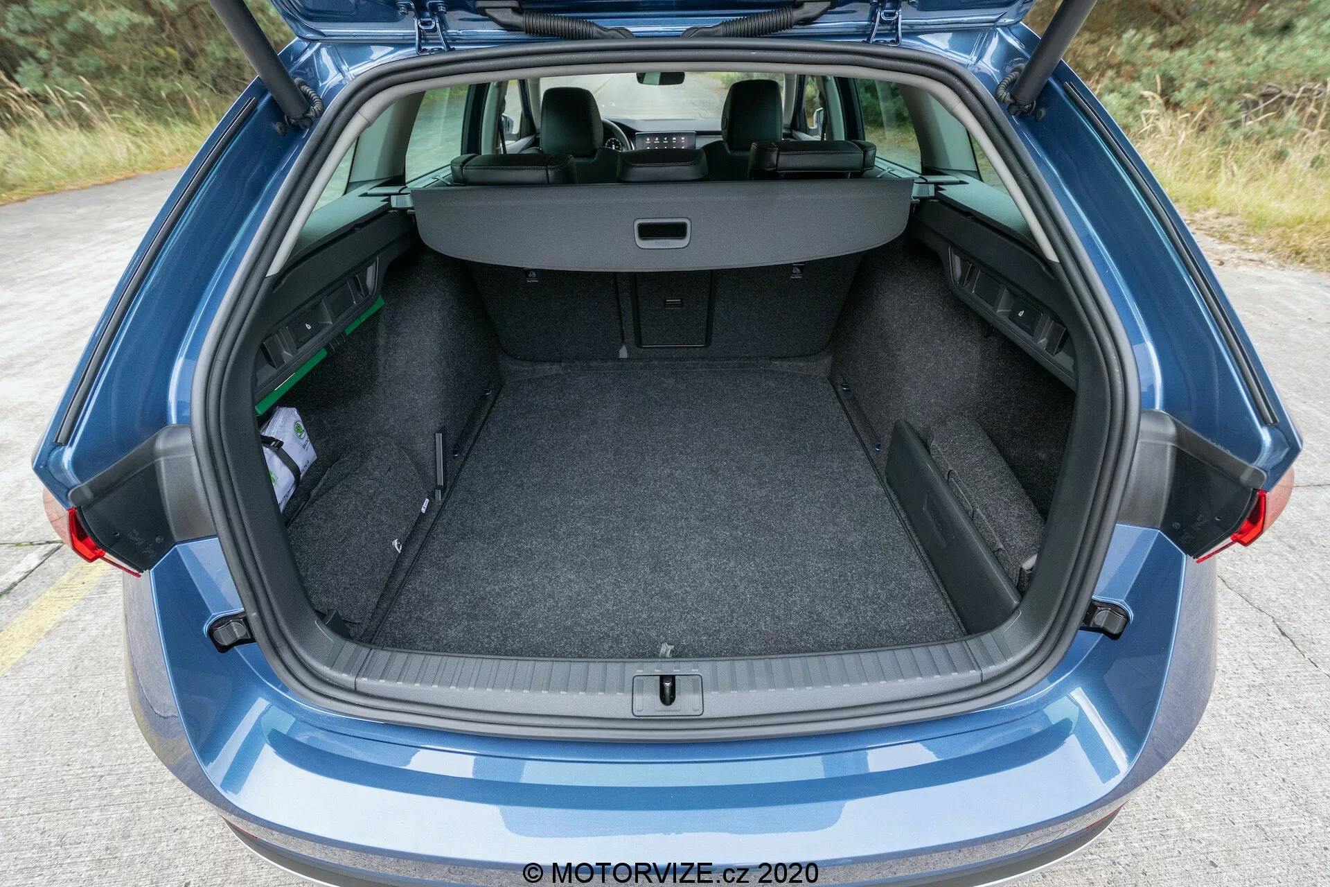 Vista de perto do porta-malas aberto de um moderno Skoda Octavia Scout 2020, com design de porta traseira totalmente aberta. A imagem captura o porta-malas do para-choque traseiro ao para-brisa traseiro, incluindo parte do teto. A área de carga acarpetada apresenta um piso de carga plano e um pequeno compartimento de armazenamento à esquerda. Os assentos traseiros estão em uma posição normal com uma cobertura retrátil estendida, ocultando os itens no porta-malas.