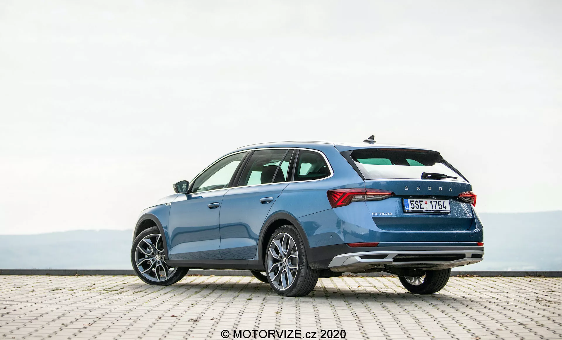 Uma imagem detalhada de um Skoda Octavia Scout 2020 azul, estacionado em uma superfície de azulejos, capturada de uma perspectiva de três quartos traseira. A fotografia destaca o perfil elegante do veículo, o design distinto do porta-malas e o lado direito. As características apresentadas incluem luzes traseiras horizontais, uma linha de caráter que sugere movimento, para-choque traseiro com refletores possivelmente abrigando sensores de estacionamento, sistema de escape duplo e rodas de liga leve com múltiplos raios. A imagem sugere um interior espaçoso, características avançadas de segurança e elementos de design contemporâneo ideais para entusiastas da tecnologia automotiva.