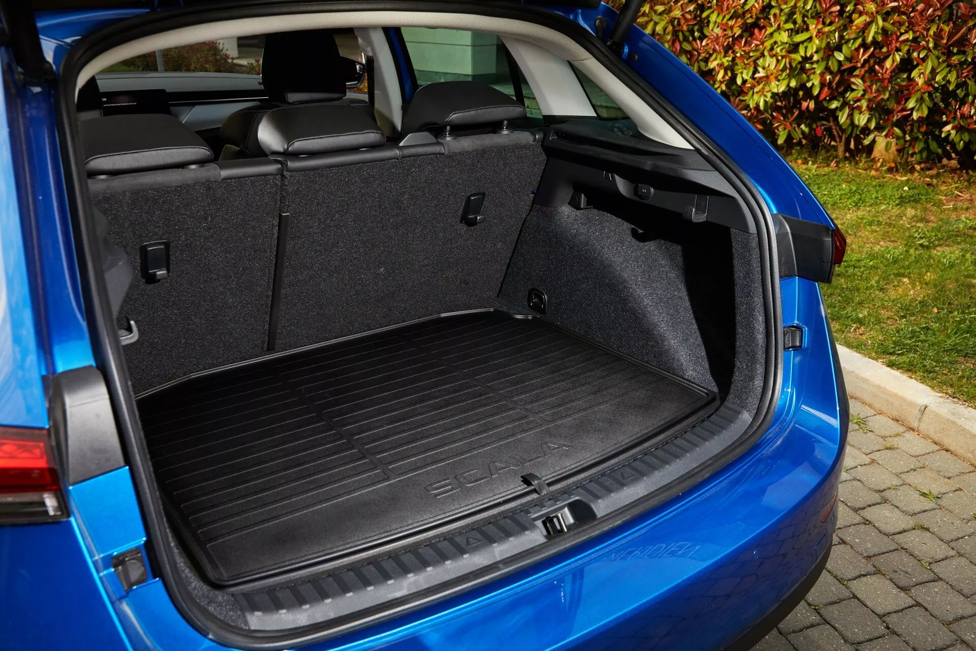Área de carga do Skoda Scala 2019 de um ângulo traseiro de três quartos, destacando o espaço do porta-malas, mecanismo da tampa do porta-malas, revestimento interno e funcionalidades como o assento traseiro rebatível 60/40, ganchos para carga e uma tomada de 12V