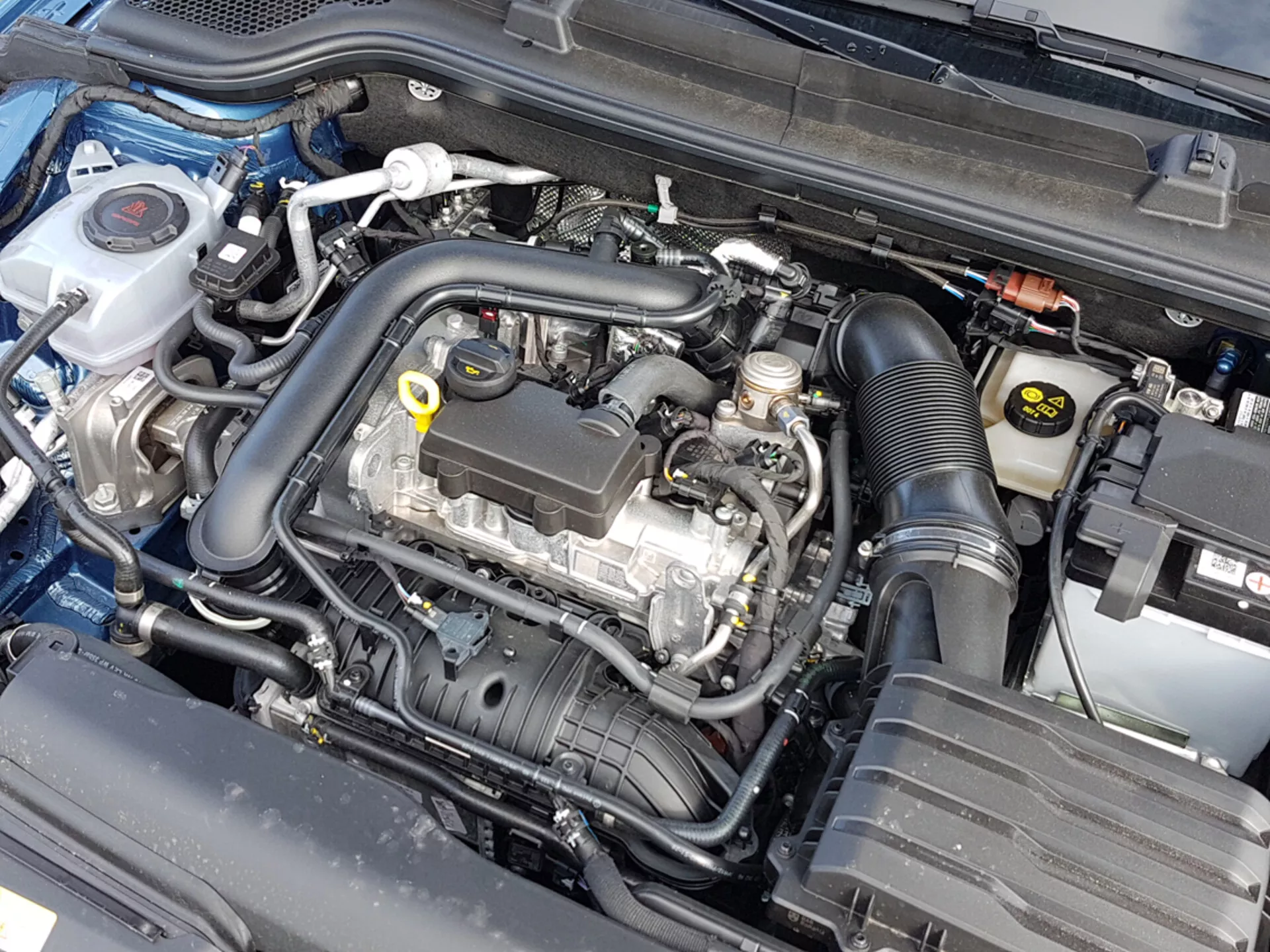 Vista detalhada do compartimento do motor de um Skoda Scala 2019 com o capô aberto, mostrando o bloco do motor e vários componentes como o recipiente de líquido de arrefecimento, a caixa do filtro de ar e a bateria, indicativo de engenharia automotiva e atenção à manutenção.