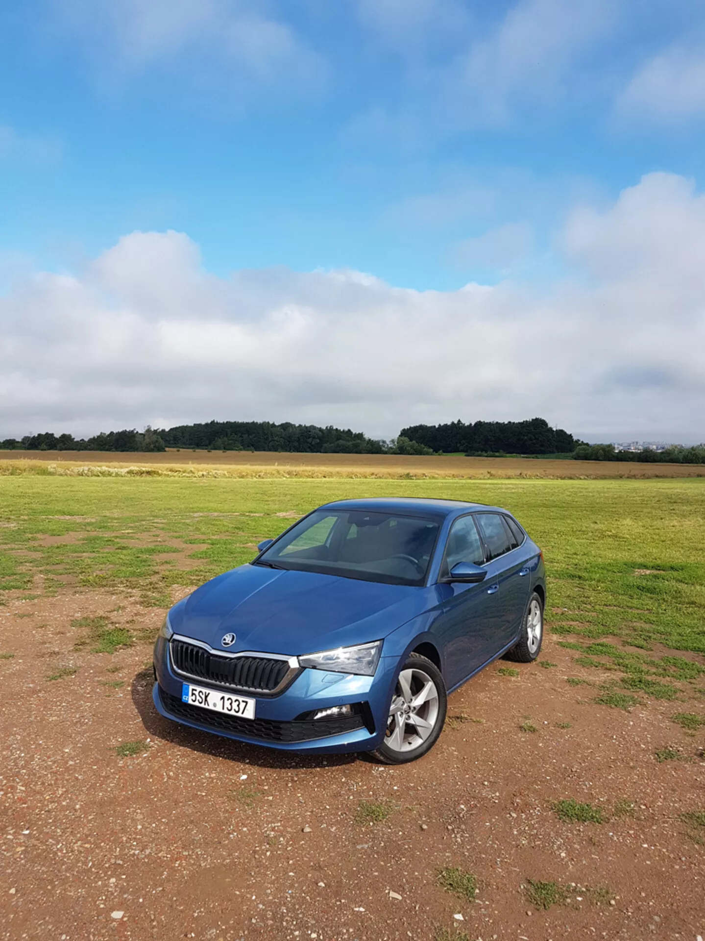 Sedan Skoda Scala 2019 azul, estacionado na grama com céu nublado, mostrando grade frontal, faróis e perfil lateral do passageiro com fundo de campo