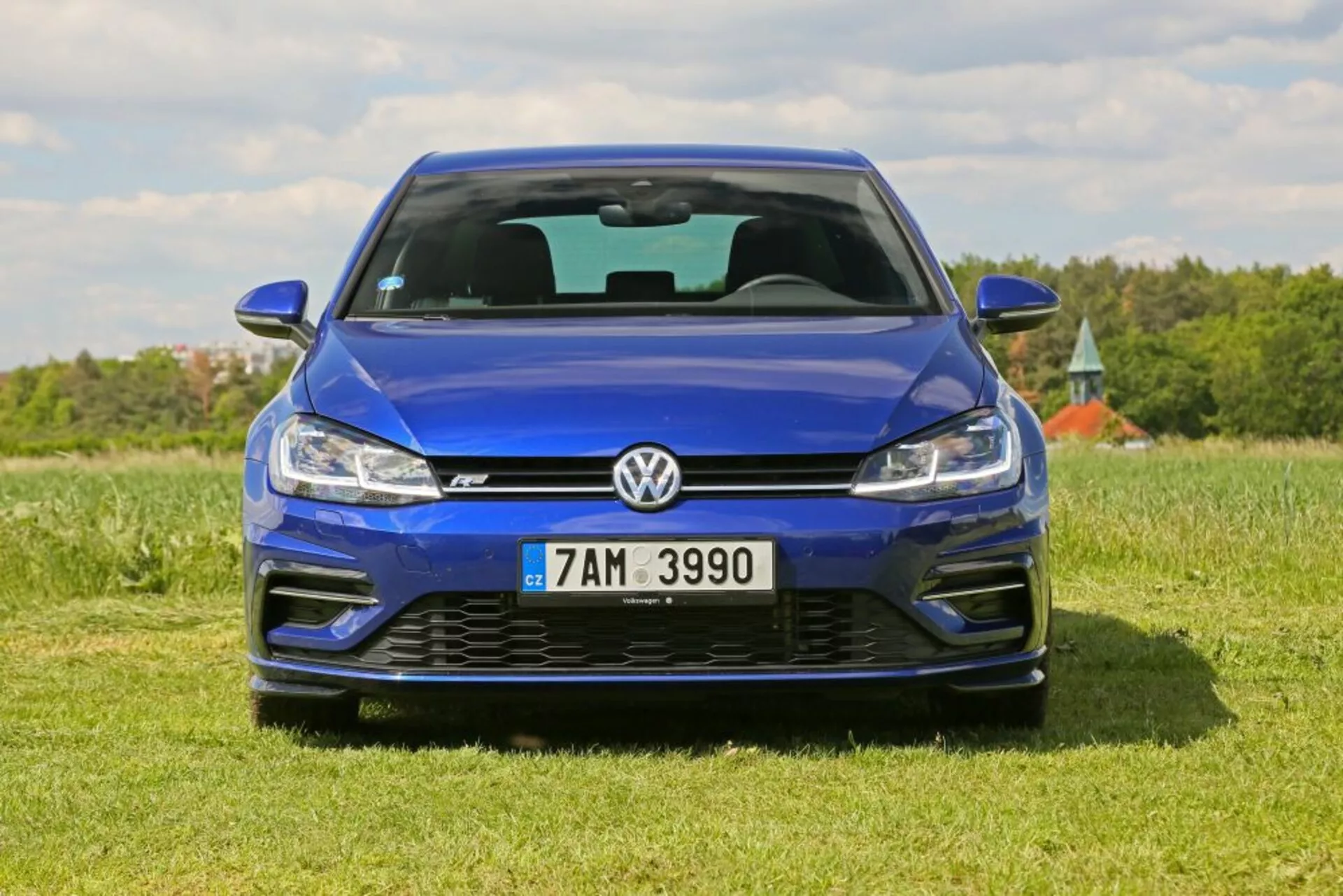 Vista frontal de um Volkswagen Golf azul estacionado em um campo gramado sob um céu claro, destacando seu acabamento metálico, design da grade, tecnologia de faróis LED e parachoque dianteiro esportivo incluindo entradas de ar e emblema.