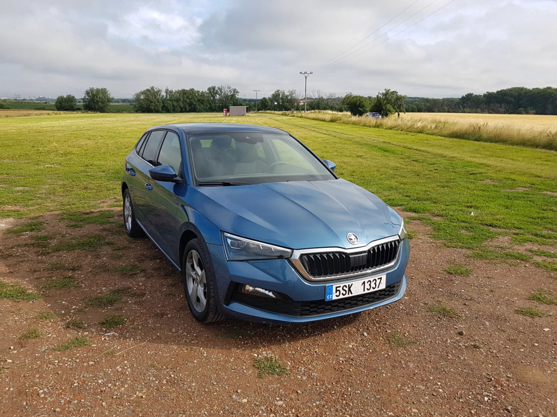 Skoda Scala azul de 2019 visto de um ângulo frontal de três quartos destacando design, faróis de LED, grade cromada, rodas de liga leve de múltiplos raios, sinais de mudança de direção integrados nos espelhos laterais e vidros com insulfilm, posicionado sobre cascalho com um campo ao fundo sob céus nublados.
