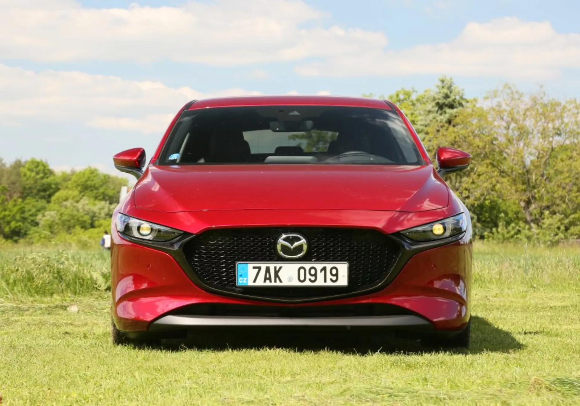 Veículo Mazda 2019 em vermelho, exibindo uma vista frontal com grade saliente apresentando o emblema da Mazda, faróis de LED e design do capô contornado. Posicionado ao ar livre na grama com árvores e um céu azul ao fundo.