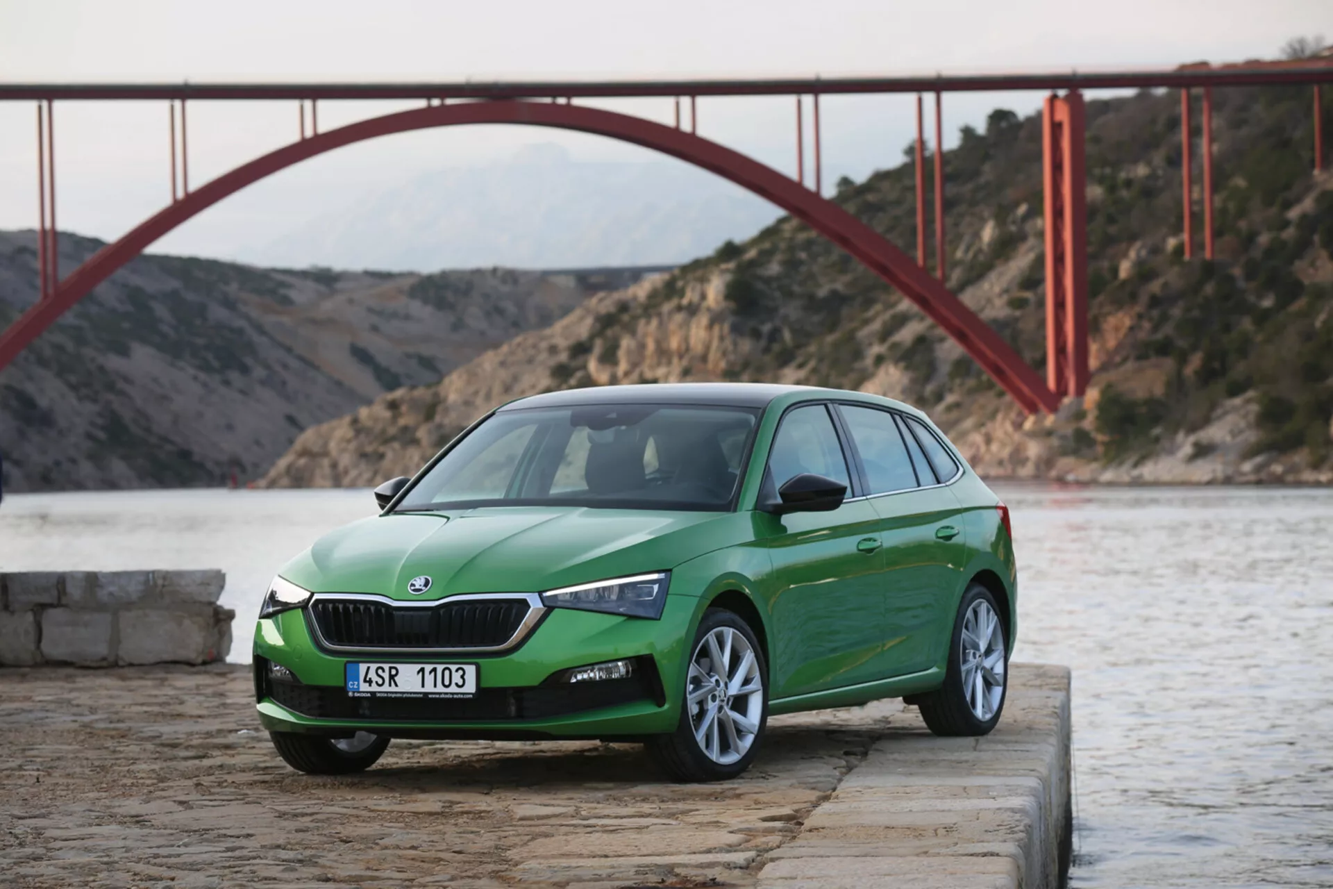 Hatchback verde de quatro portas Skoda Scala 2019 em cais de concreto perto da água com grade visível, faróis de LED, para-choque dianteiro e rodas de liga leve, contra um pano de fundo de ponte cênica