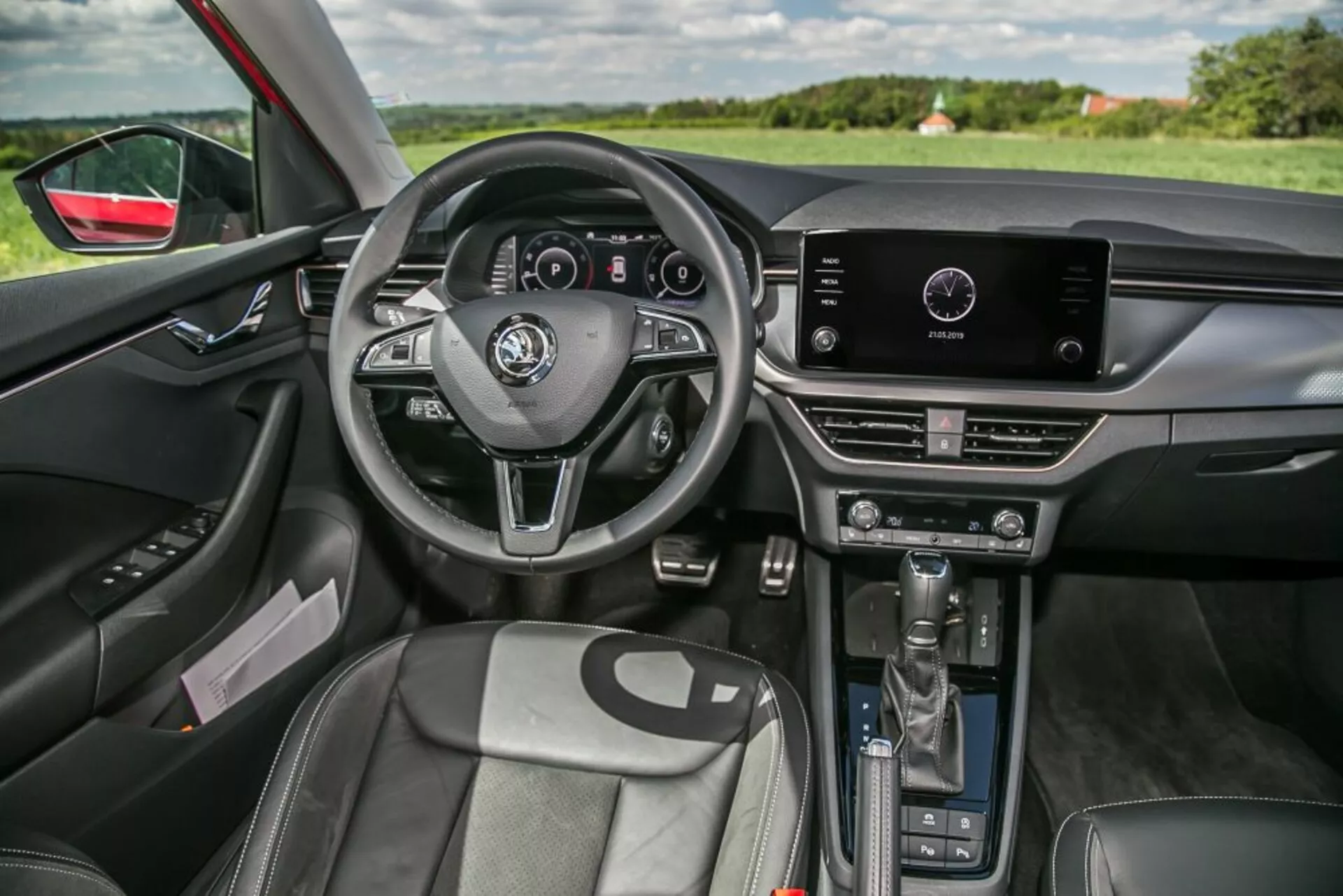 Vista interior do Skoda Scala 2019 do lado do passageiro frontal apresentando volante com três raios com controles integrados e cluster de instrumentos digital, sistema de infotainment touchscreen no topo do console central com controles manuais do clima abaixo, alavanca de câmbio automática, assentos frontais reforçados com estofamento tipo couro, painel da porta do lado do motorista com ajustes de janelas e espelhos elétricos, iluminação ambiente interna, espelho retrovisor com atenuação automática, e visibilidade da paisagem através do para-brisa e janela do lado do motorista, destacando design ergonômico e tecnologia integrada.