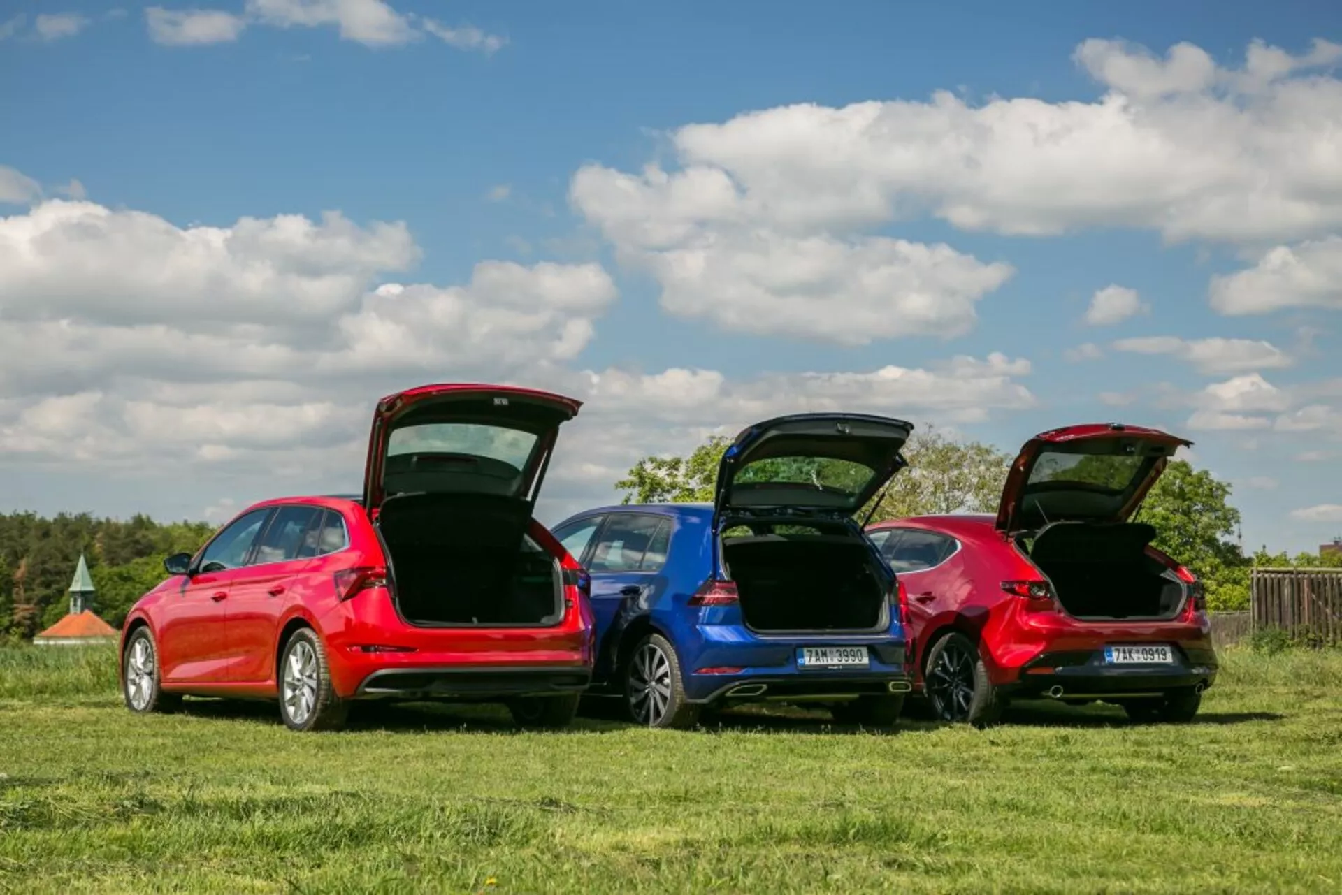 Modelos Skoda Scala 2019 em vermelho e azul estacionados em um campo gramado, exibindo perfis traseiros, bagageiros abertos para funcionalidade do espaço de carga e detalhes de design sob um céu claro.