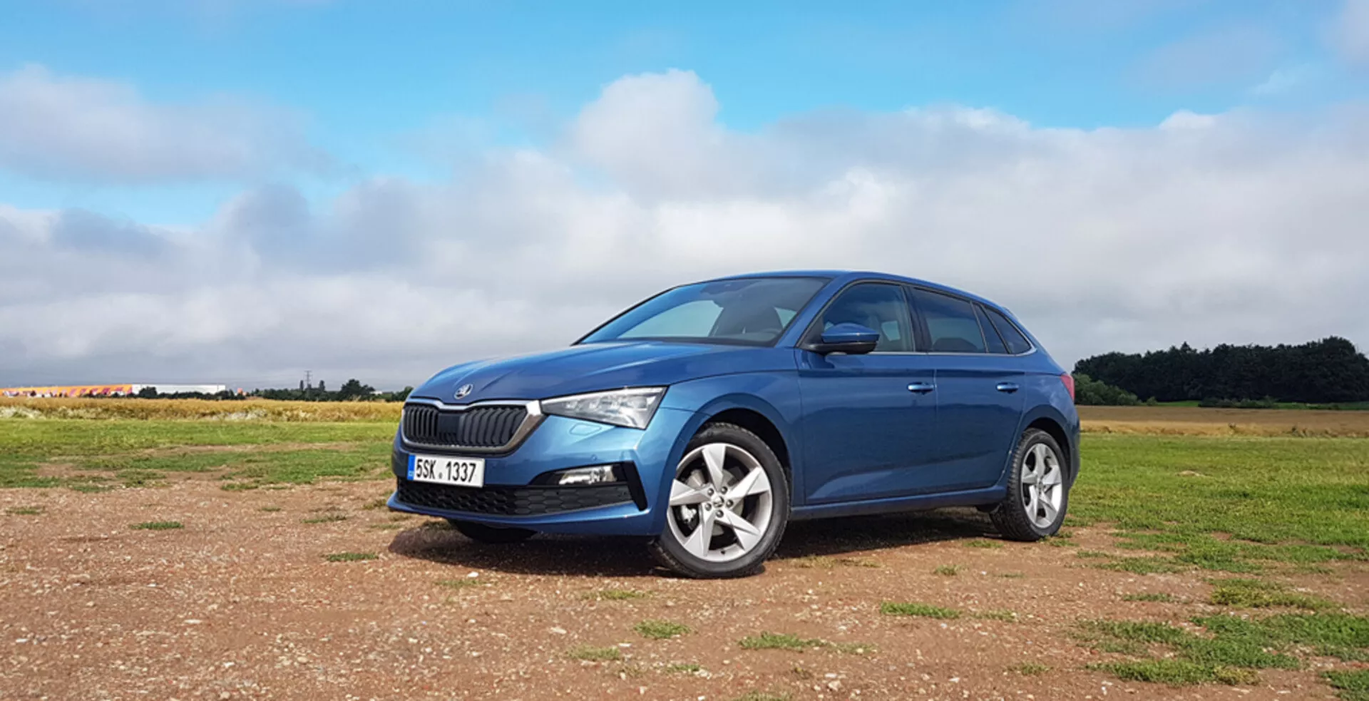 Hatchback azul Skoda Scala 2019 em superfície não pavimentada com grade frontal distintiva, faróis dianteiros LED modernos, rodas de liga leve e um perfil lateral esquerdo elegante contra um fundo de céu parcialmente nublado