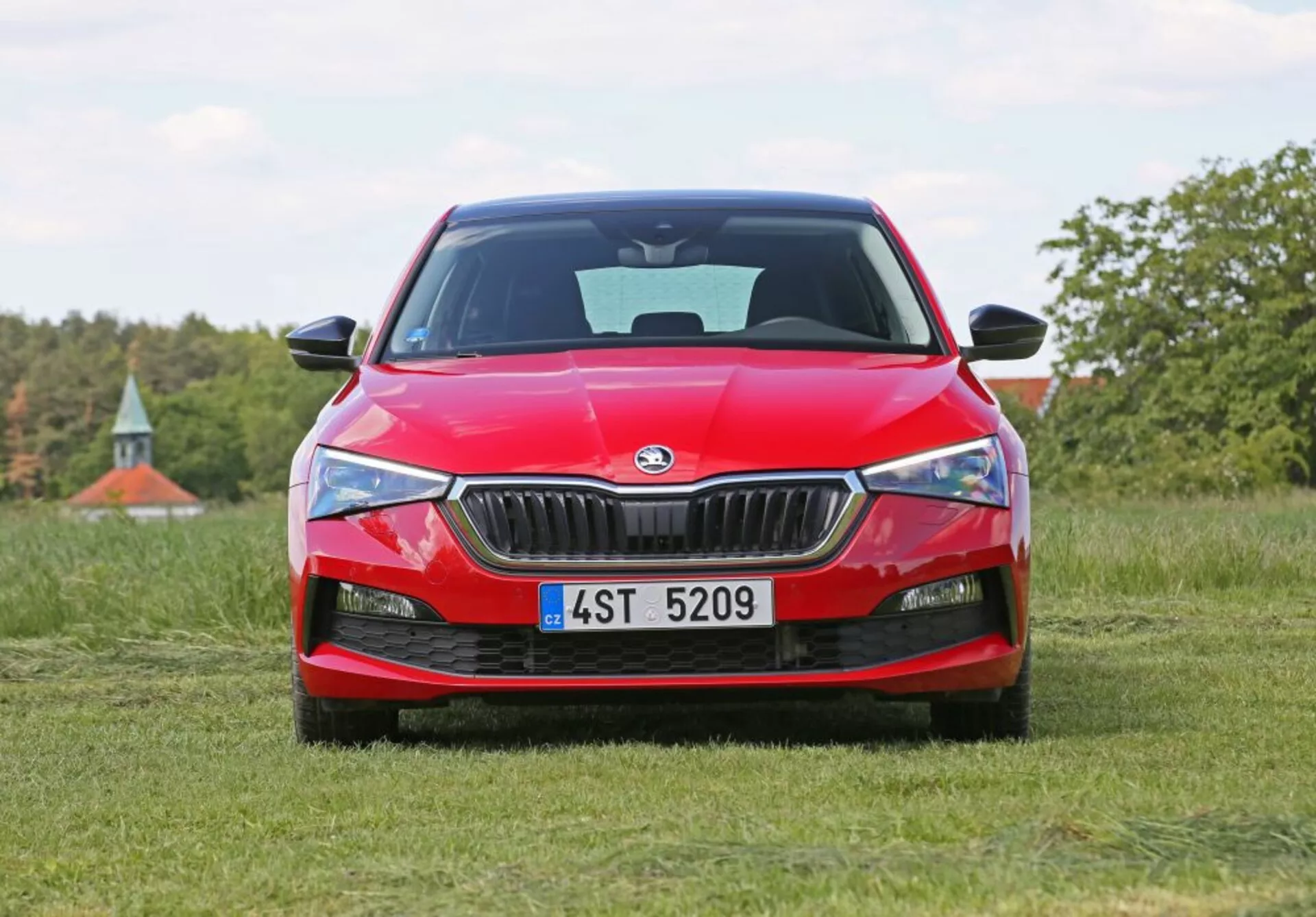 Skoda Scala 2019 em vermelho posicionado em um campo gramado com visão frontal, exibindo a grade frontal do carro, faróis aguçados e logotipo da Skoda, contra um fundo natural com árvores e um toque de céu azul.