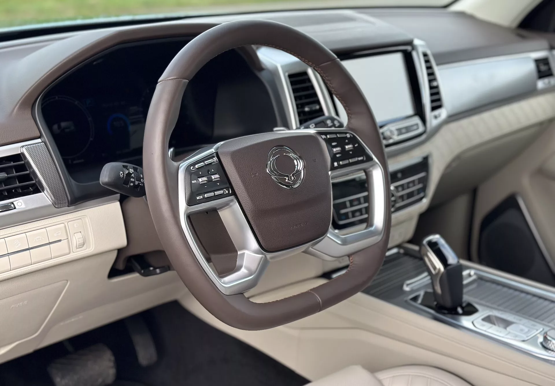 Vista interior luxuosa da SsangYong Rexton 2021 do lado do passageiro da frente, apresentando um volante multifuncional revestido a couro com acentos metálicos, painel de instrumentos digital, sistema de infotainment avançado e esquema de cores em dois tons bege e escuro.