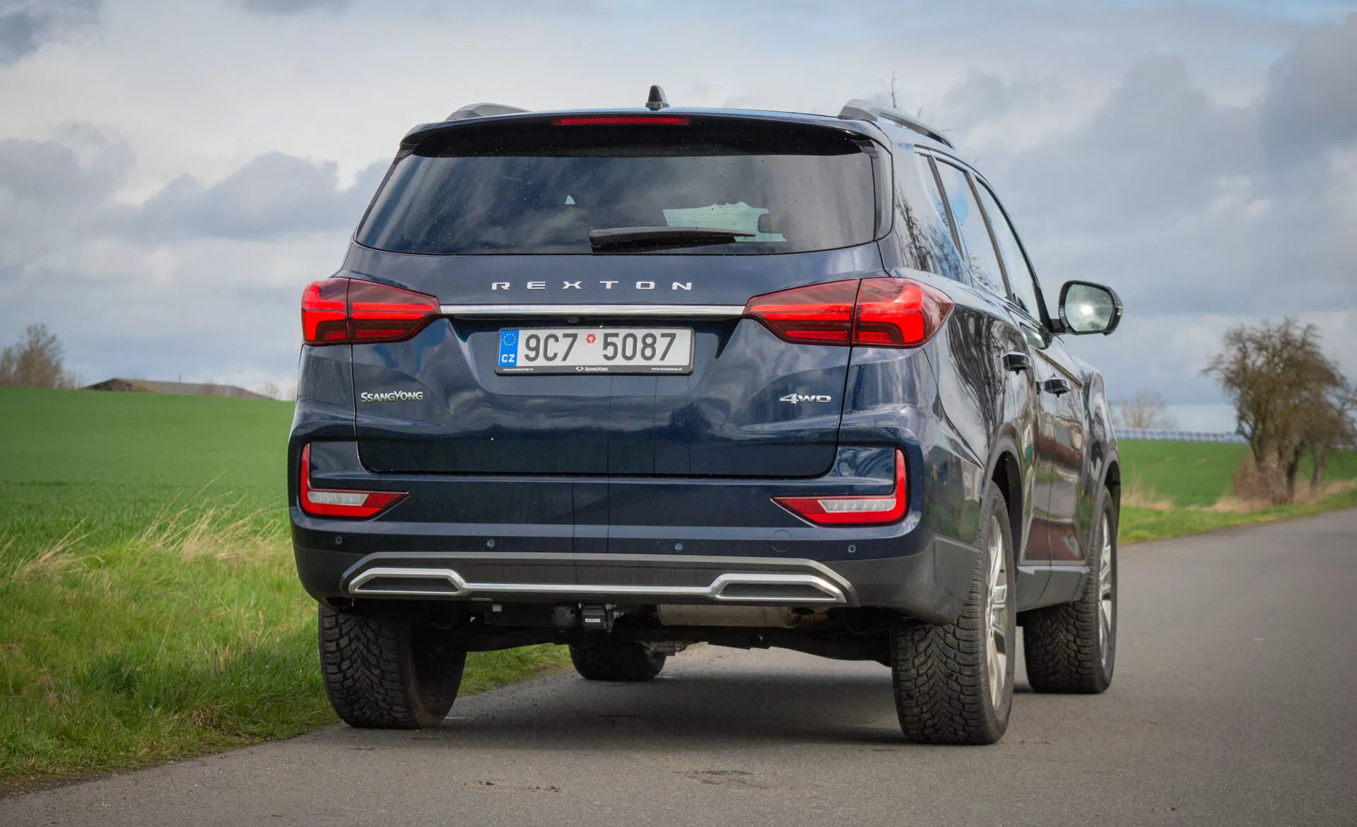 Vista traseira de um SUV azul SsangYong Rexton 2021 estacionado num caminho rural de duas vias, destacando as suas características de design como luzes traseiras, janela traseira, tubos de escape e tecnologia de segurança incluindo câmera de ré e sensores de estacionamento, sob um céu nublado.