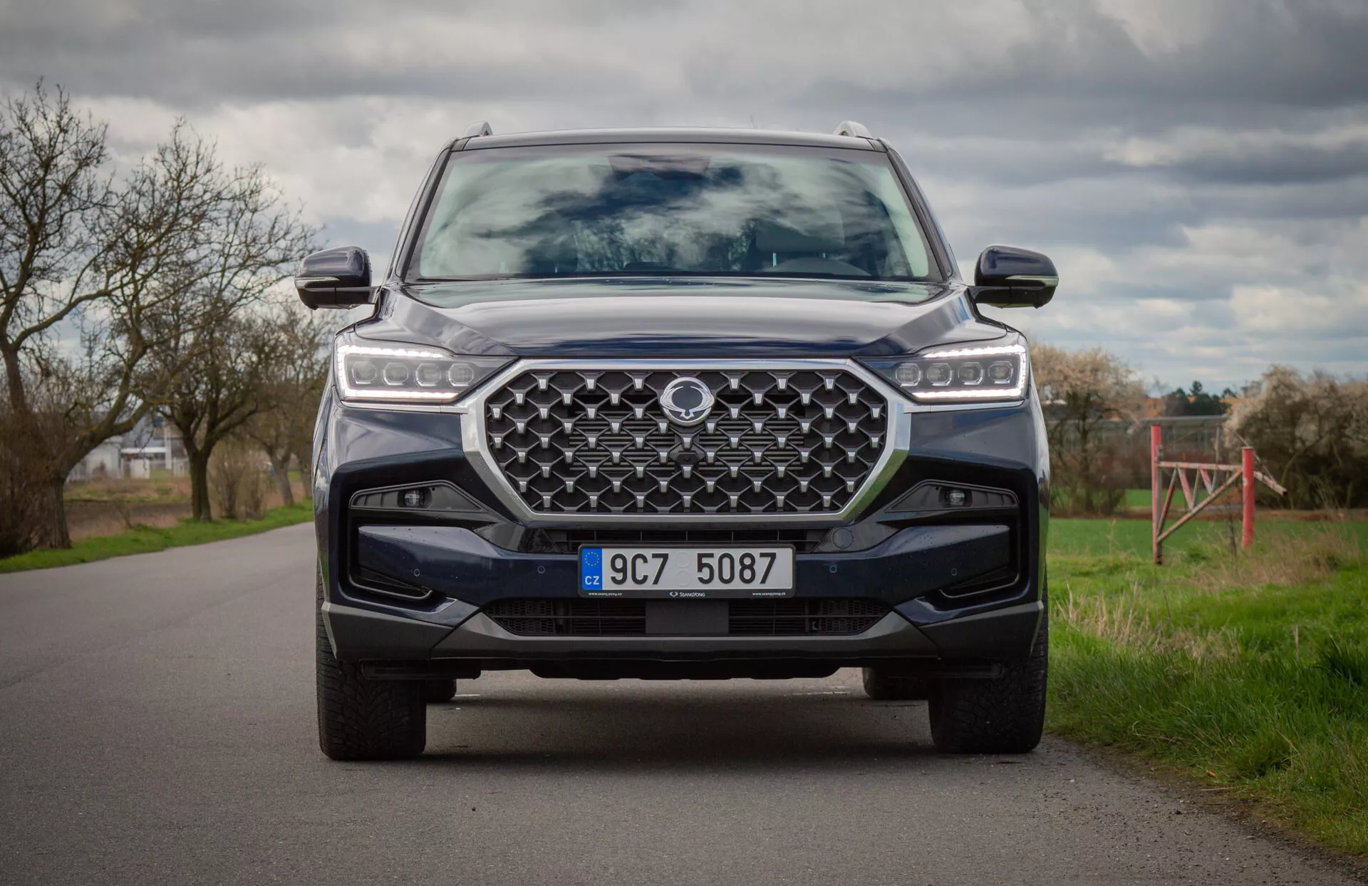 SUV moderno SsangYong Rexton 2021 em cor, capturado de uma posição ajoelhada exibindo a frente do veículo, faróis LED, grade frontal grande desenhada, para-choque dianteiro com placa de proteção e sistemas avançados de assistência ao condutor